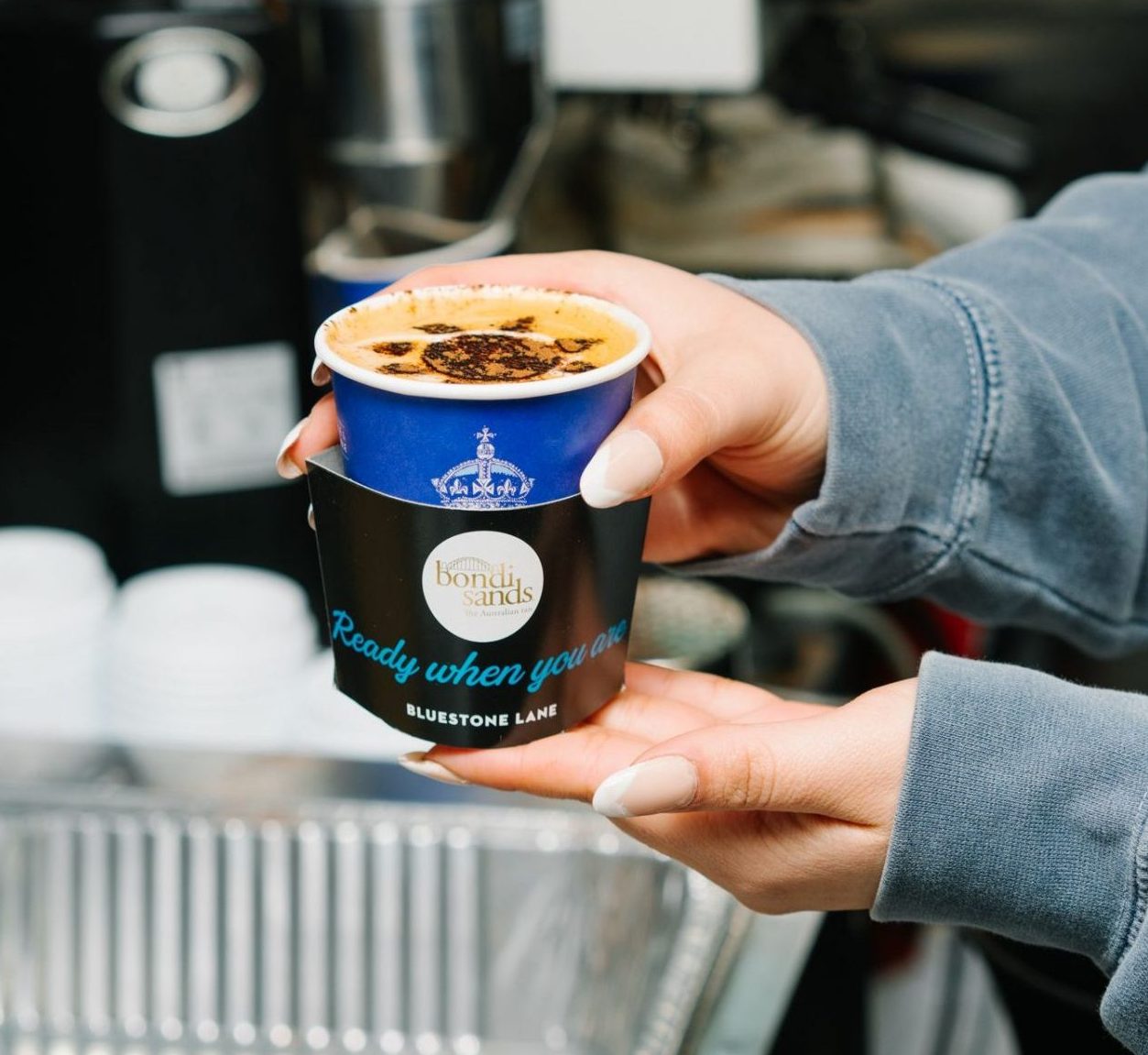 Person with manicured nails in a gray sleeve holds a blue “bondi sands” coffee cup over a sink.