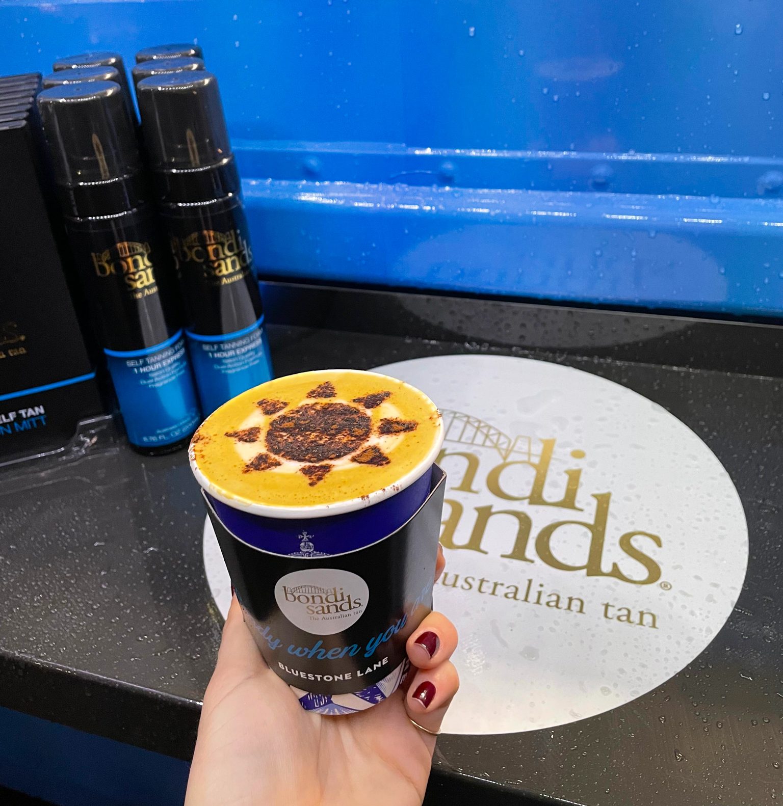Hand holding a coffee with sun-shaped froth, Bondi Sands bottles on black counter in back.