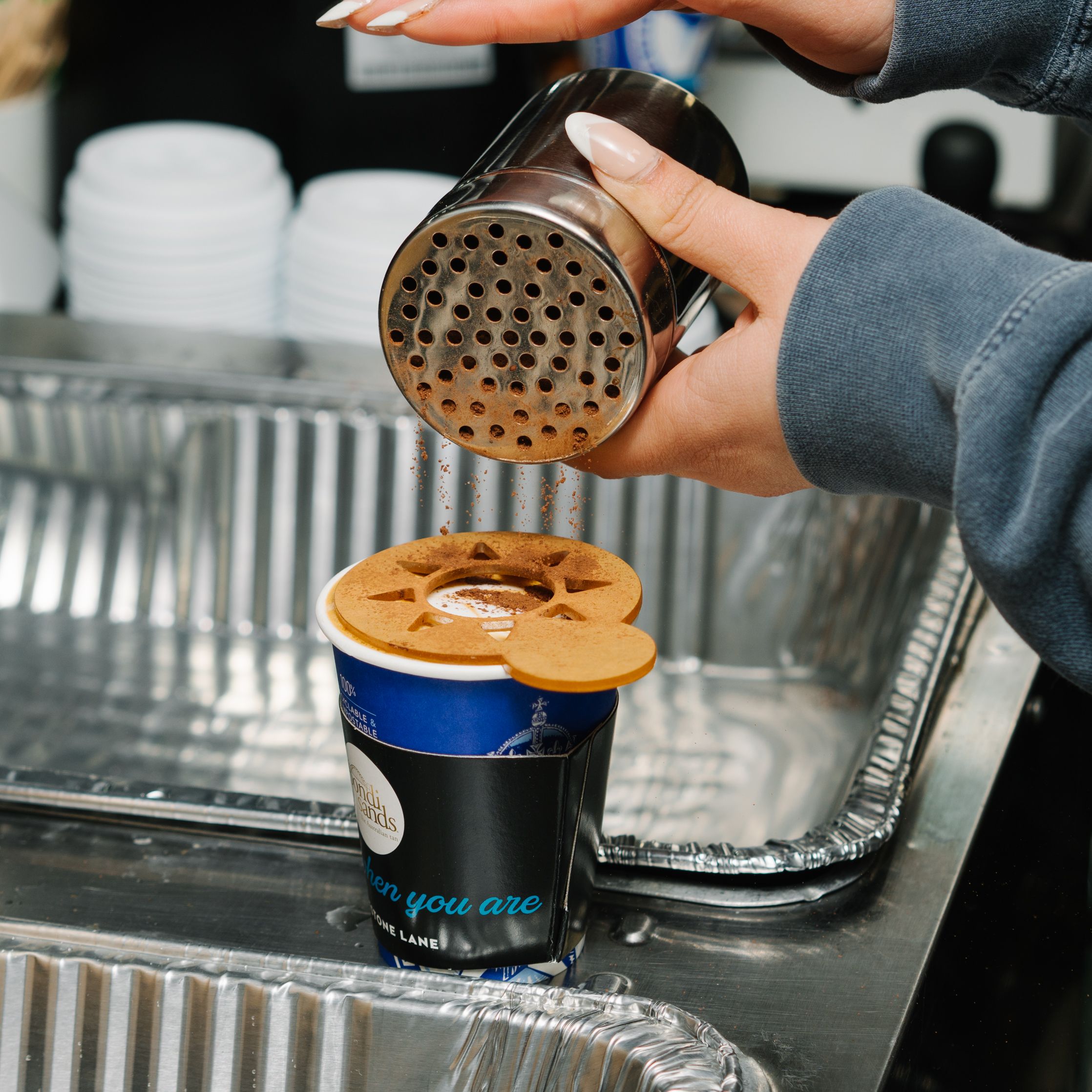 A person sprinkles cocoa on a takeaway coffee cup with a stencil, next to trays and stacked cups.