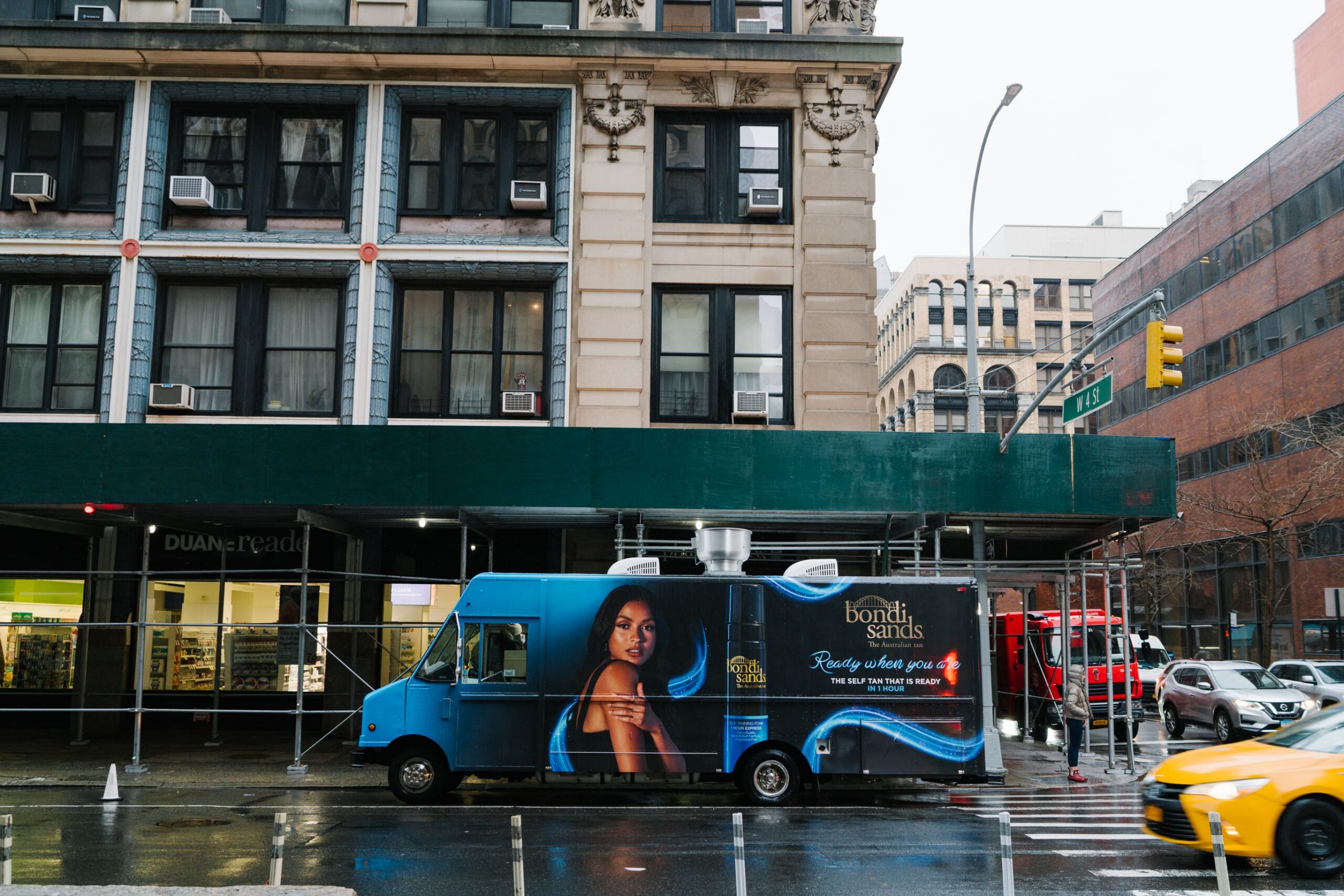Blue Bondi Sands beauty truck parked on wet city street near green scaffolding and Duane Reade.