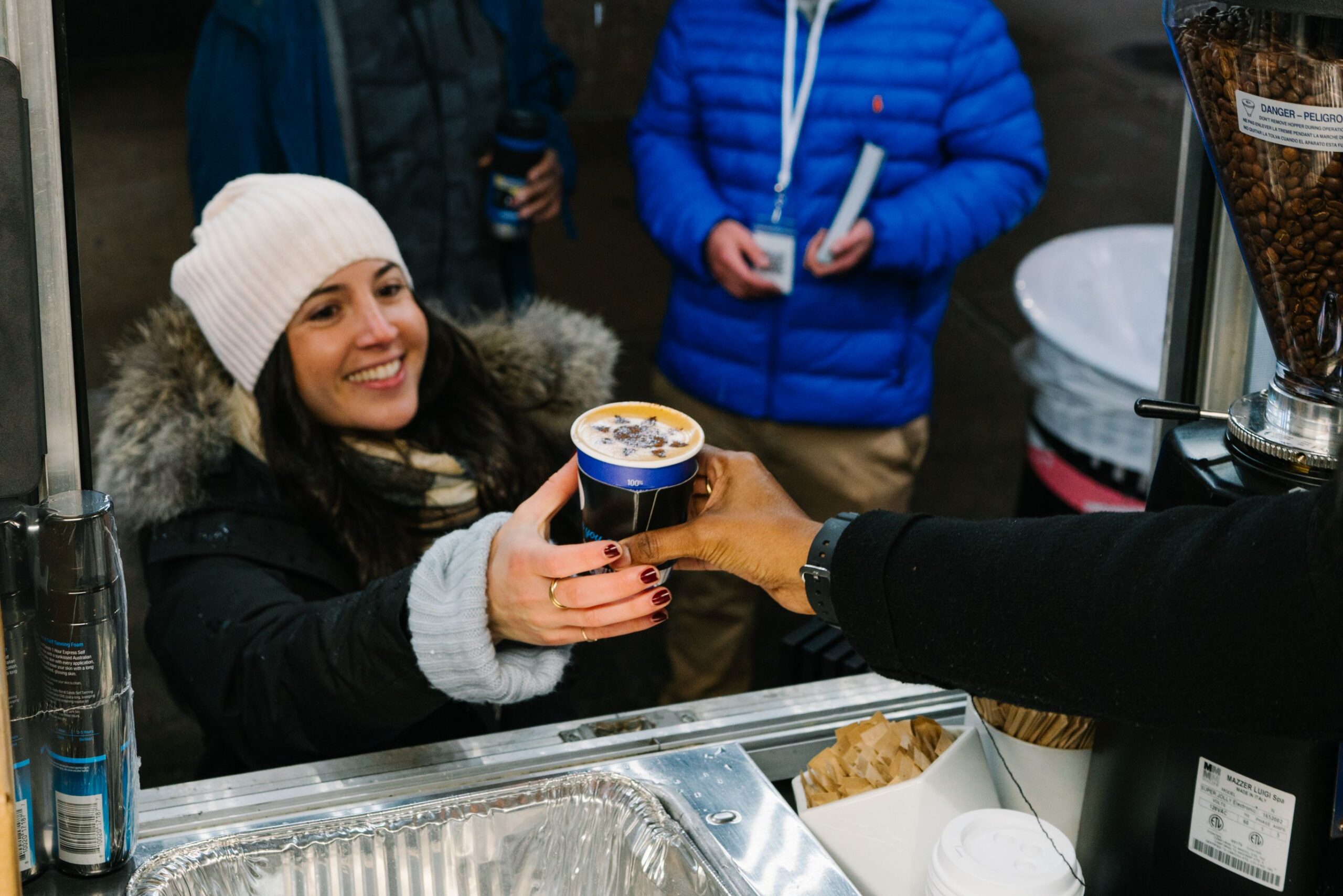 A woman in a winter coat gets coffee from a barista at an outdoor stand; others wait nearby.
