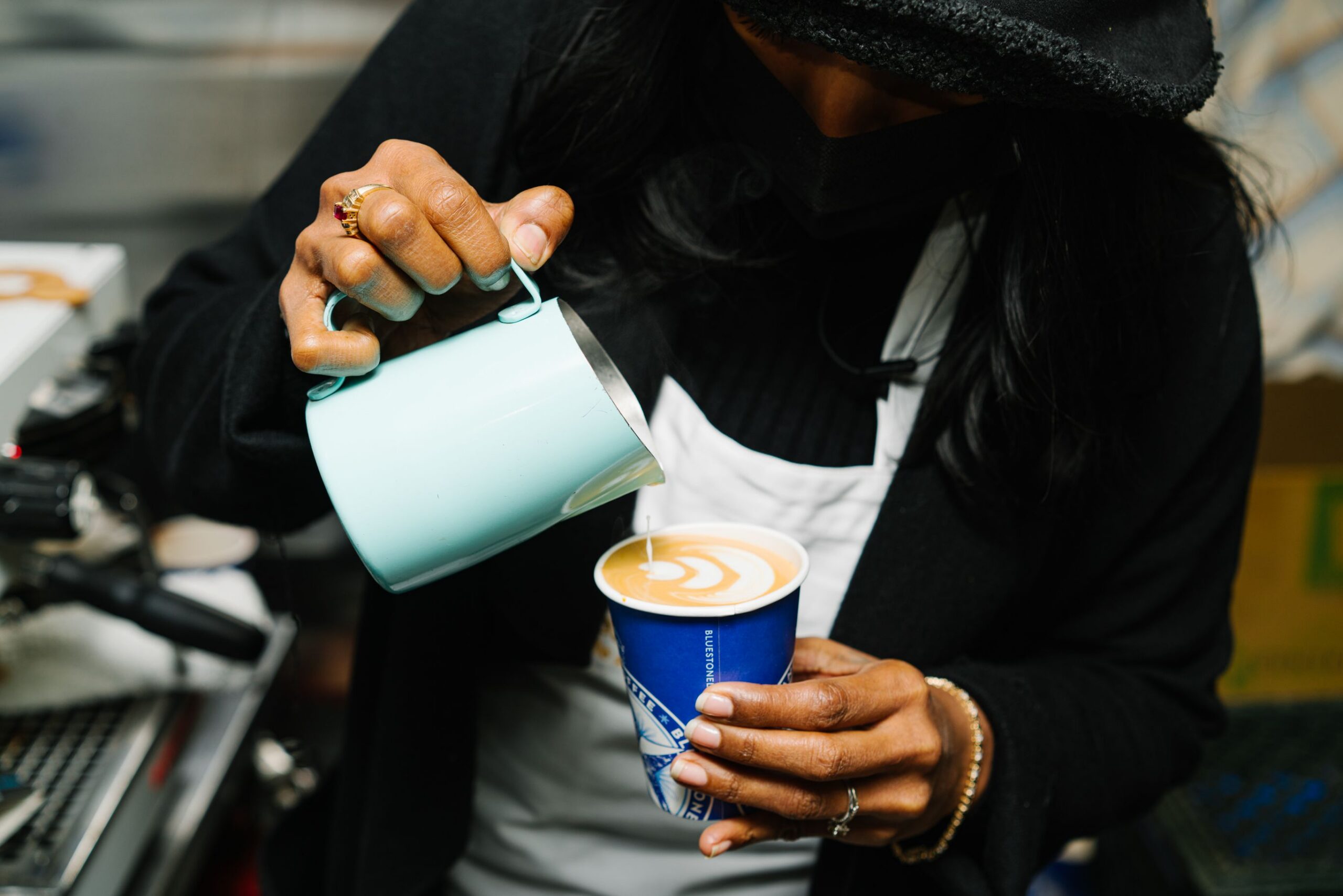 A person pours steamed milk into a blue coffee cup, making latte art. Wears black and white.