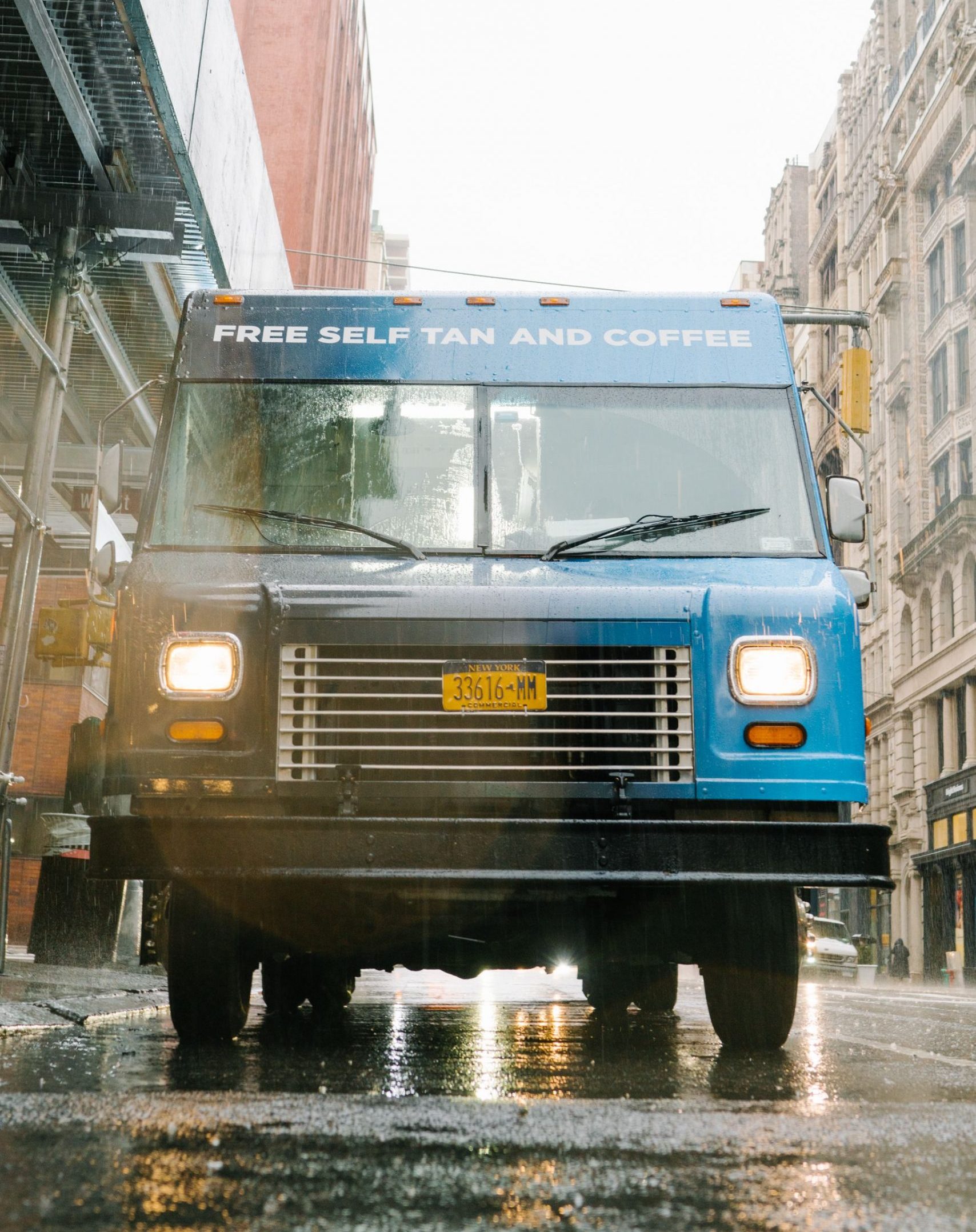 Blue delivery truck in rain offers free self tan and coffee; buildings in the background.