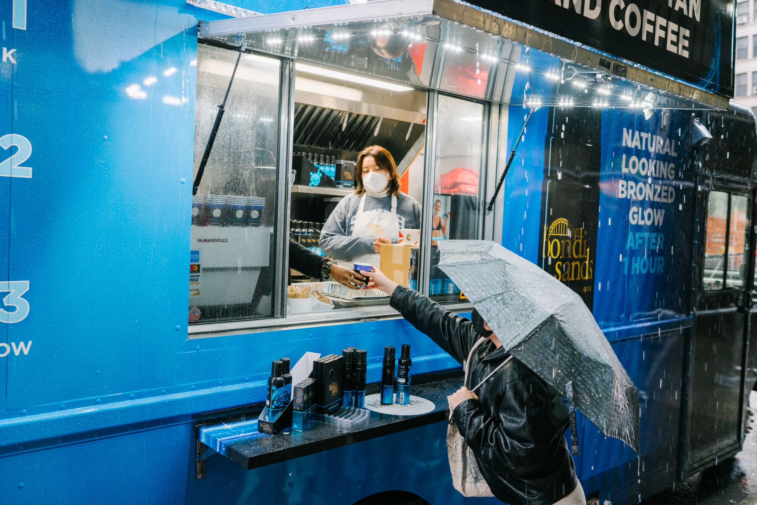 Masked woman serves a customer with an umbrella at blue pop-up food truck on a rainy day.