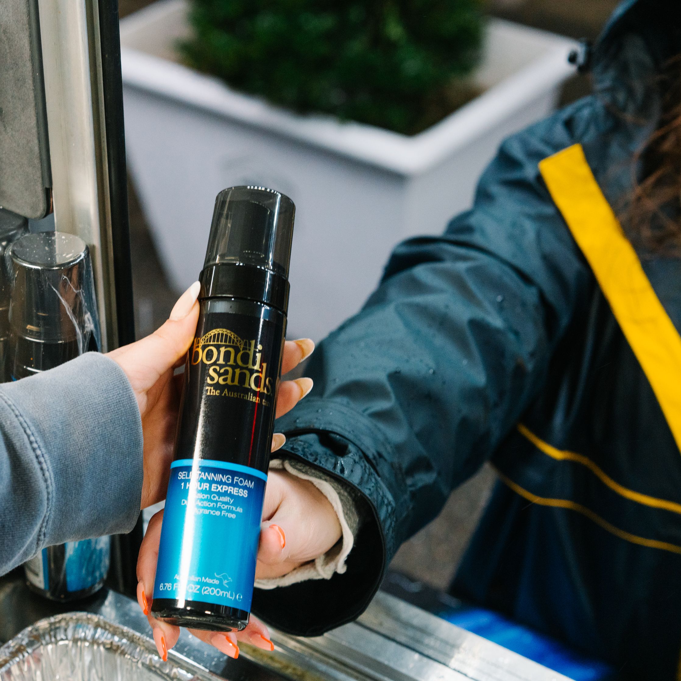 A person in a blue and yellow jacket hands someone Bondi Sands self-tanning foam at a counter.