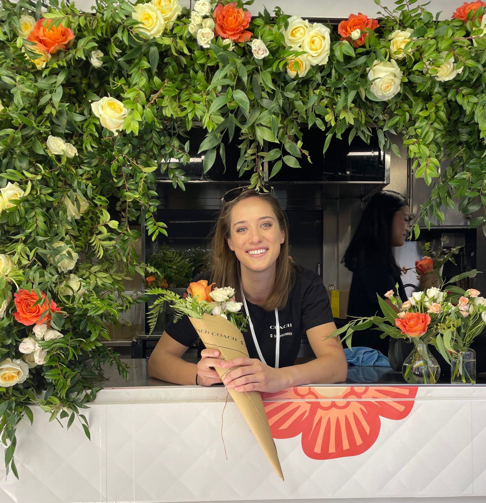 Woman smiles at a flower-filled counter, holding a bouquet with more flowers beside her.