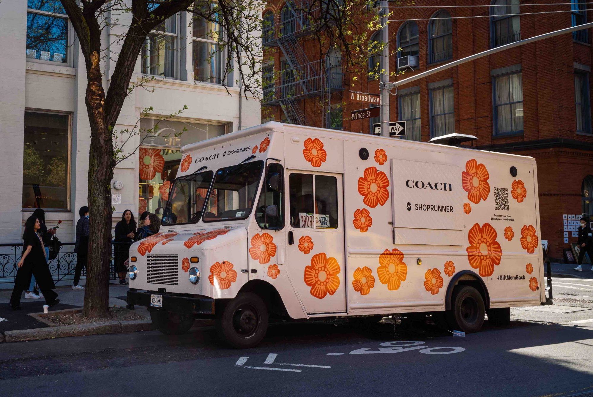 A white Coach truck with orange flowers is parked on a city street for a Mother's Day pop-up.