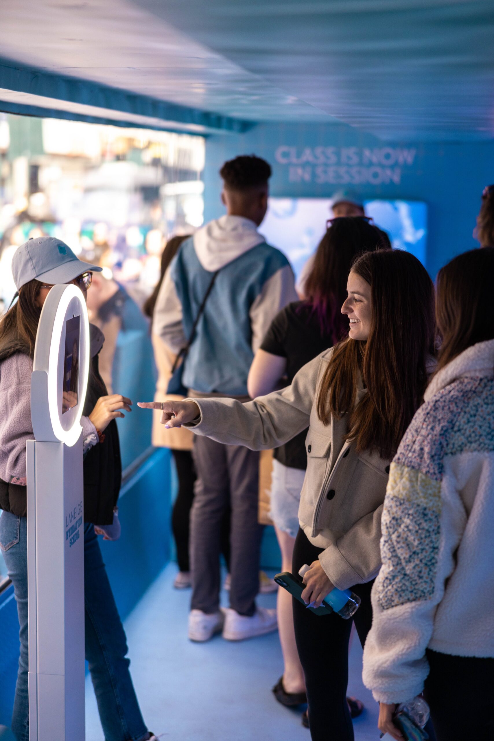 A young woman points at a Laneige selfie booth in a blue room as people wait in line behind her.