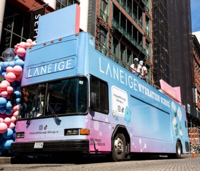 A blue and purple Laneige Hydration School bus is parked on a city street with people on top.