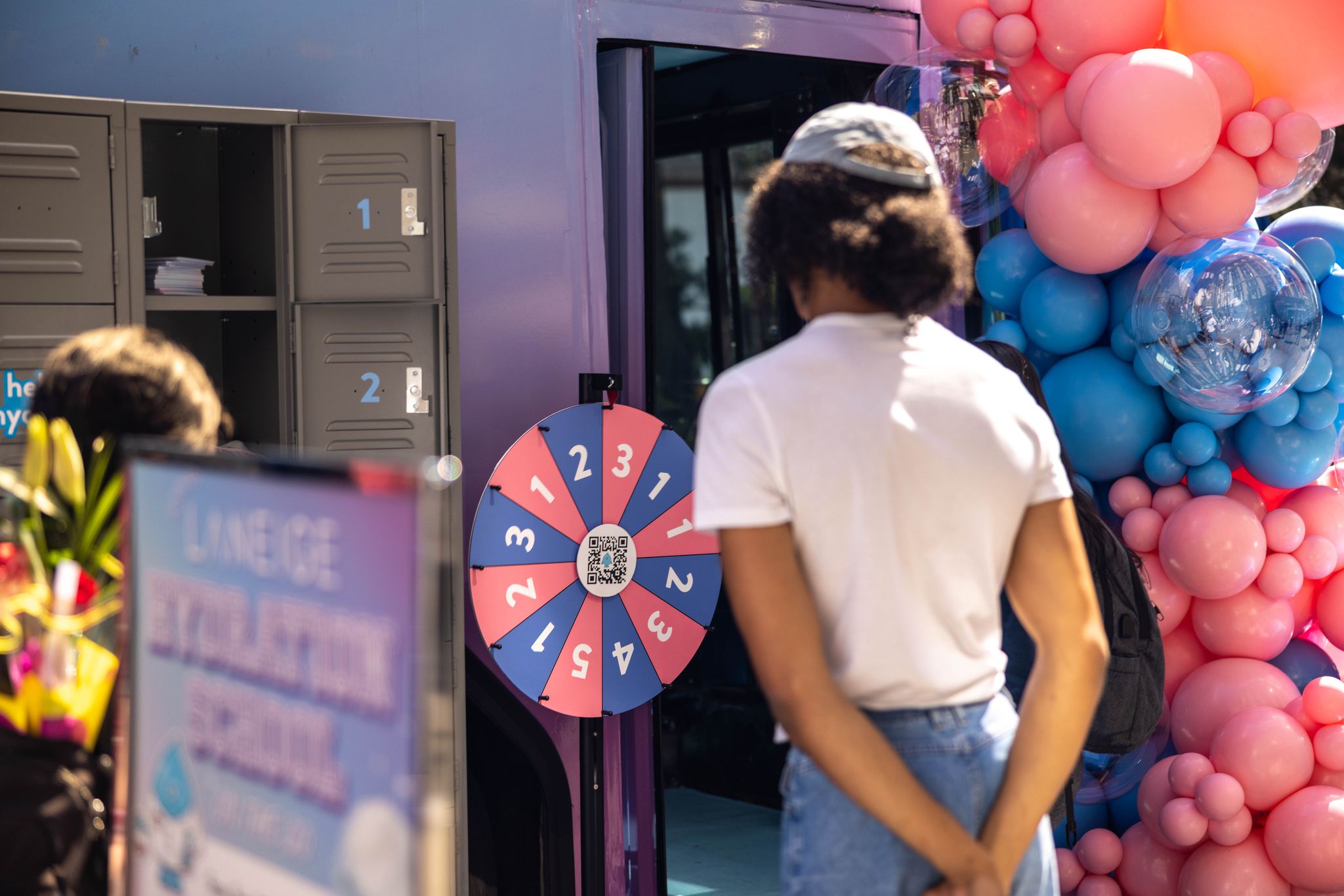 Person stands by a prize wheel, lockers, and pink and blue balloons at a Laneige pop-up event.