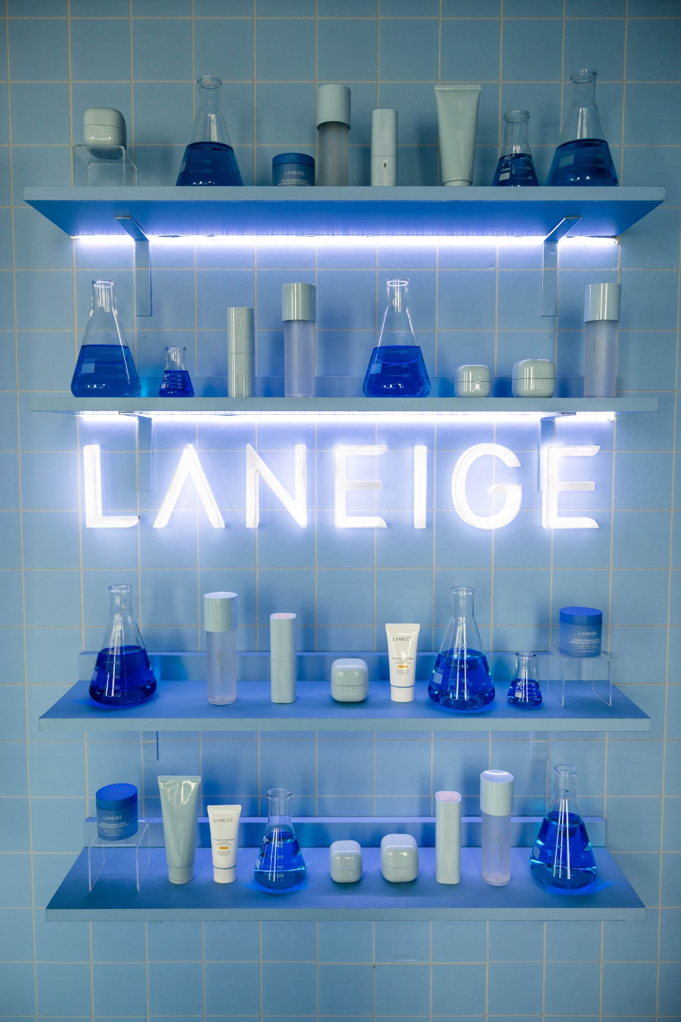 Laneige skincare and blue flasks on glass shelves with a neon LANEIGE sign on a blue-tiled wall.