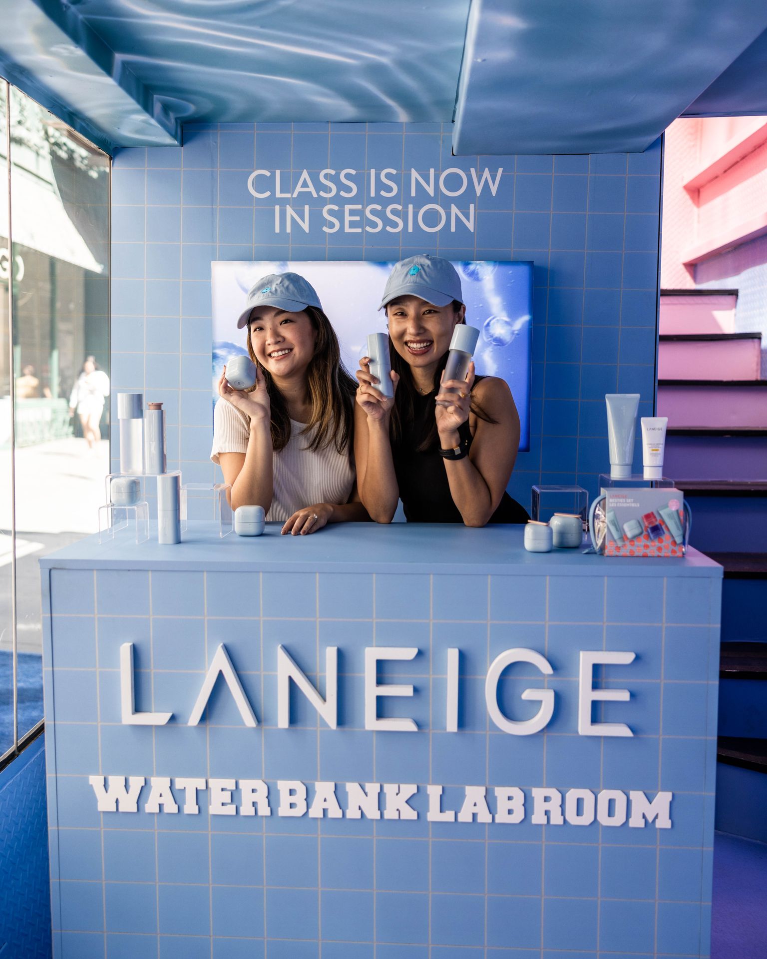 Two women in blue hats hold Laneige products at a booth with skincare items on the counter.