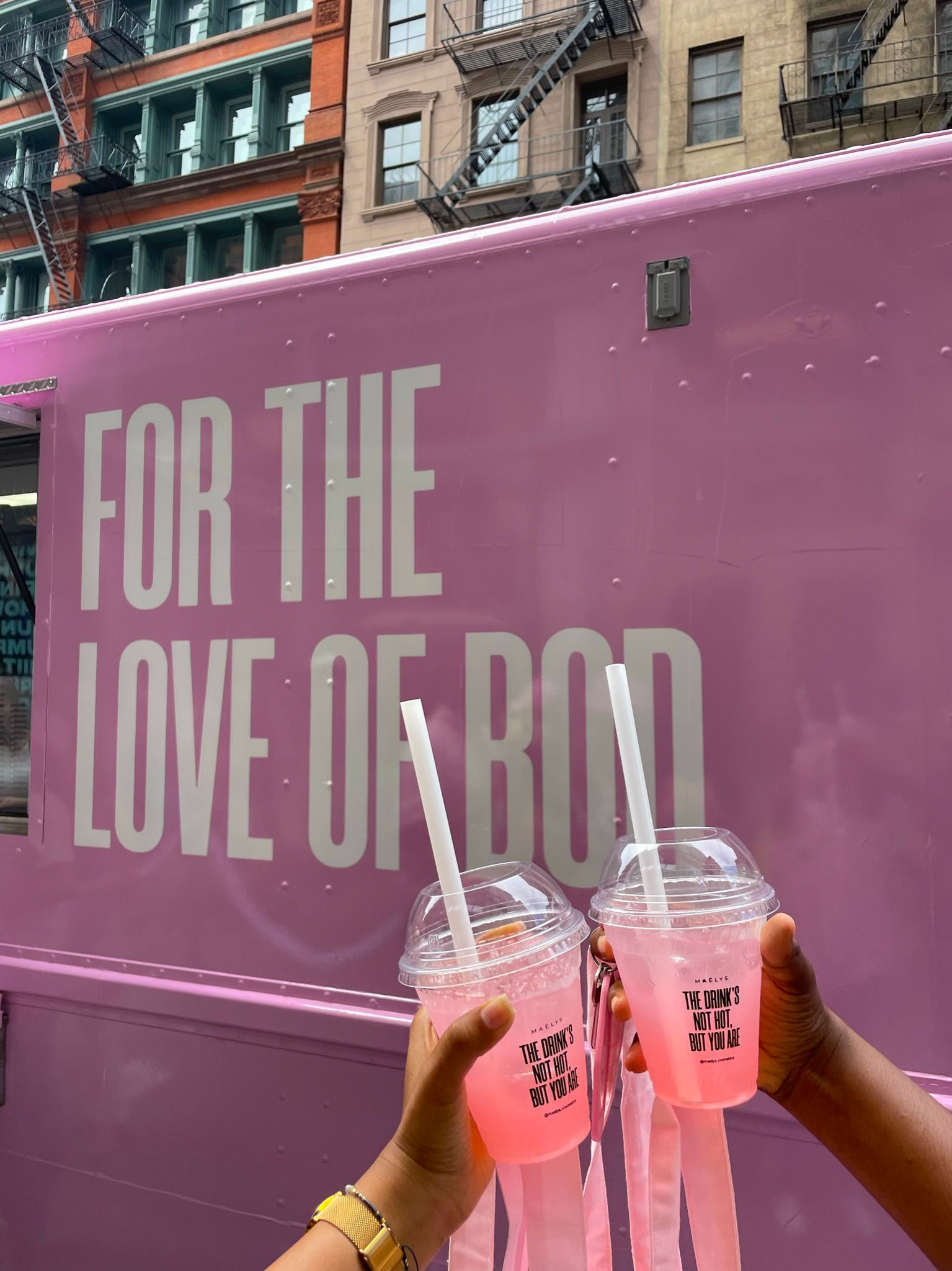 Two hands hold pink drinks with straws in front of a pink truck reading FOR THE LOVE OF GOD.