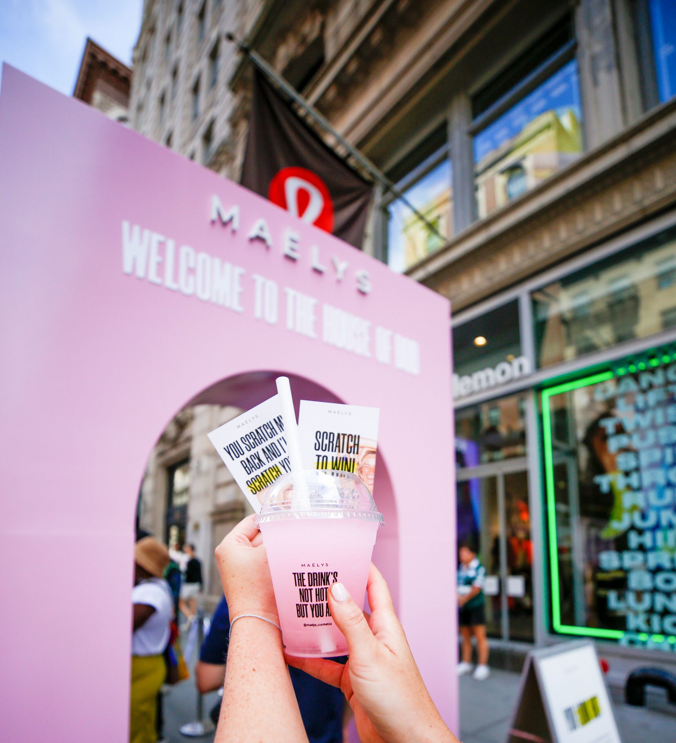 Person holds a pink drink and two Scratch to Win cards by a MAËLYS archway on a city street.