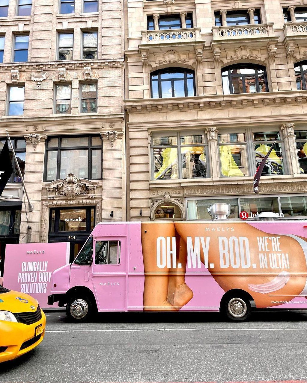 Pink Maëlys truck with large text parked on city street near ornate buildings and a yellow taxi.
