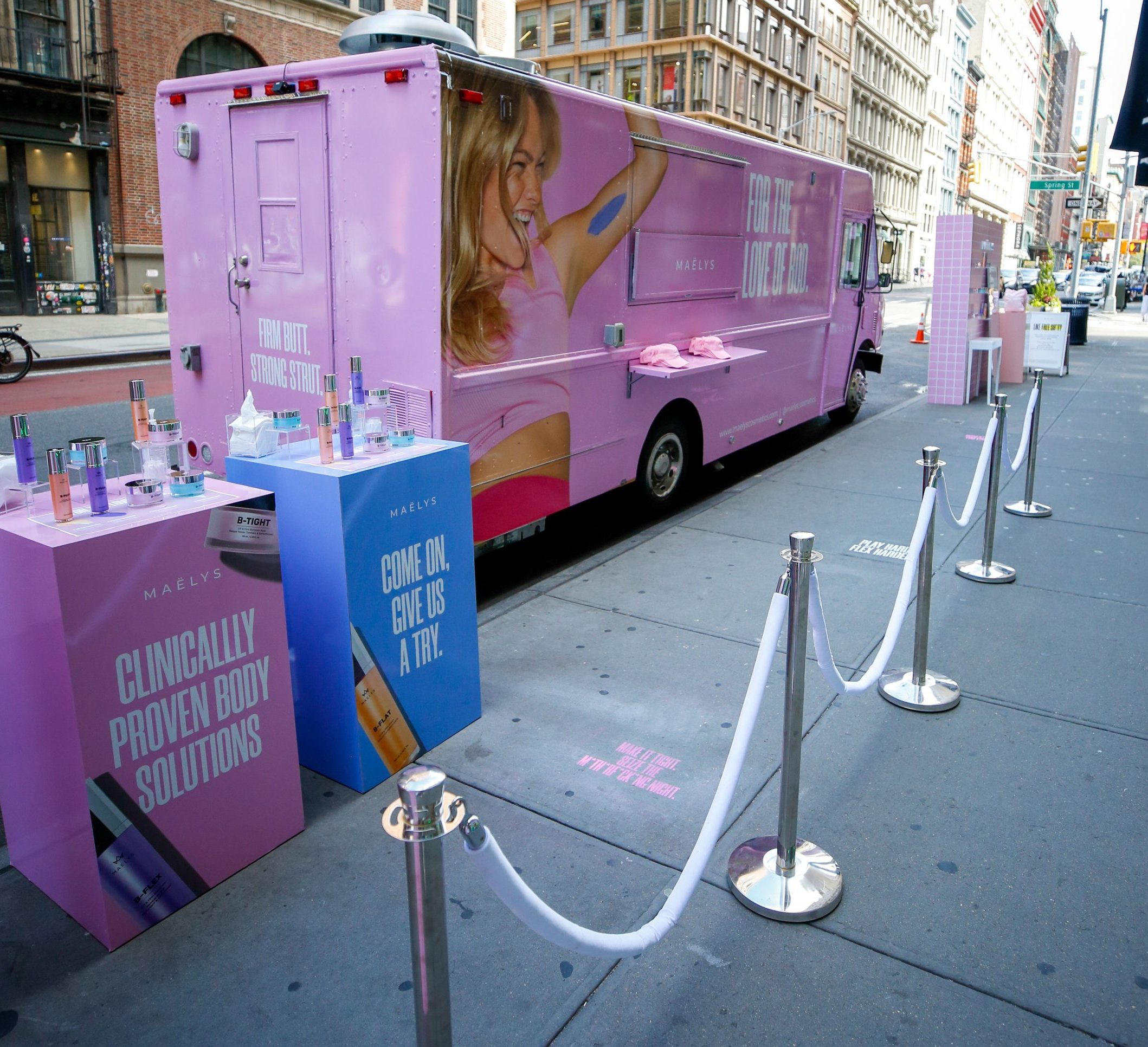 Pink MAËLYS truck parked on city street with branded stands displaying skincare products nearby.
