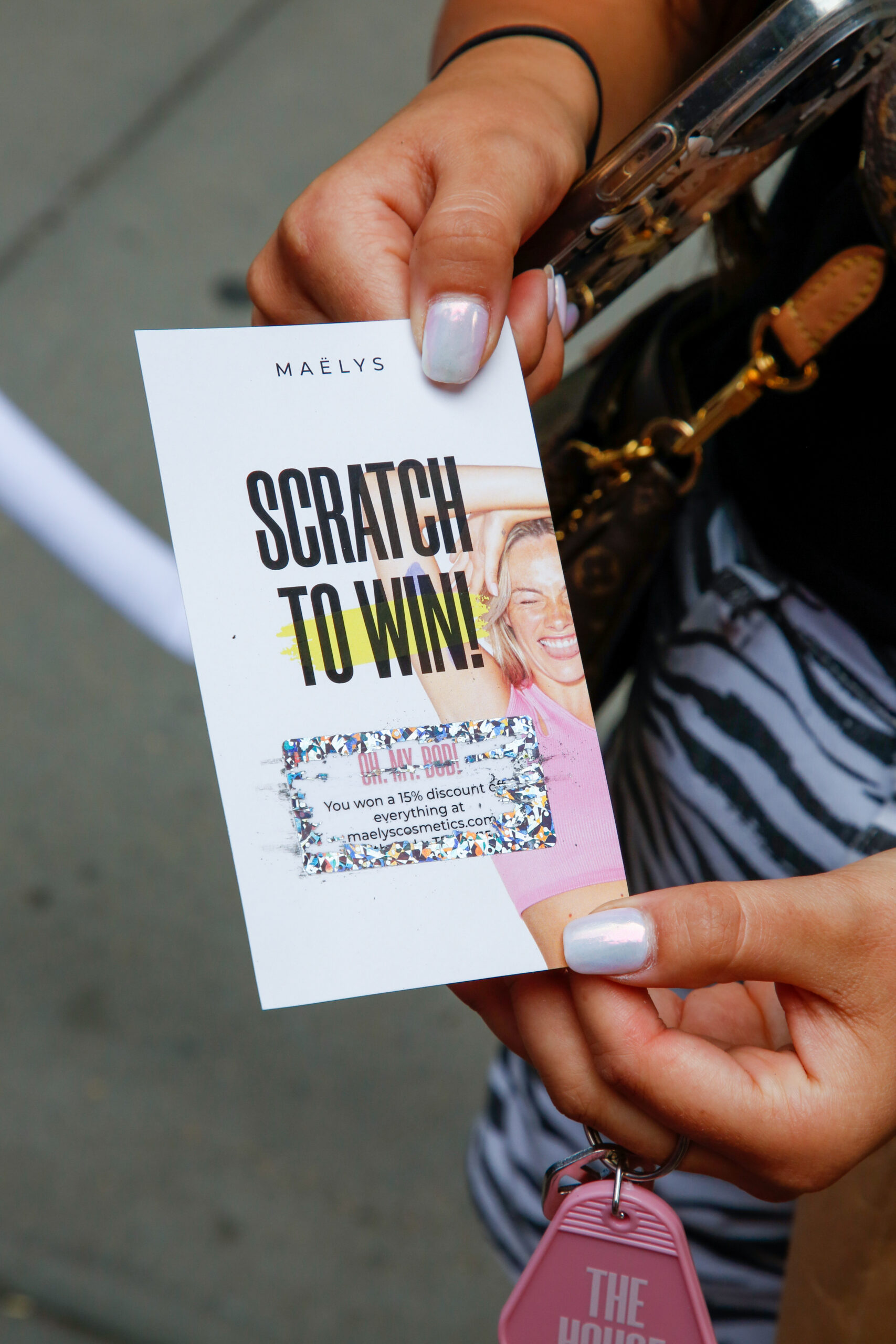 Hand with pale nail polish holds a MAËLYS scratch card showing 15% off; purse and keys behind.