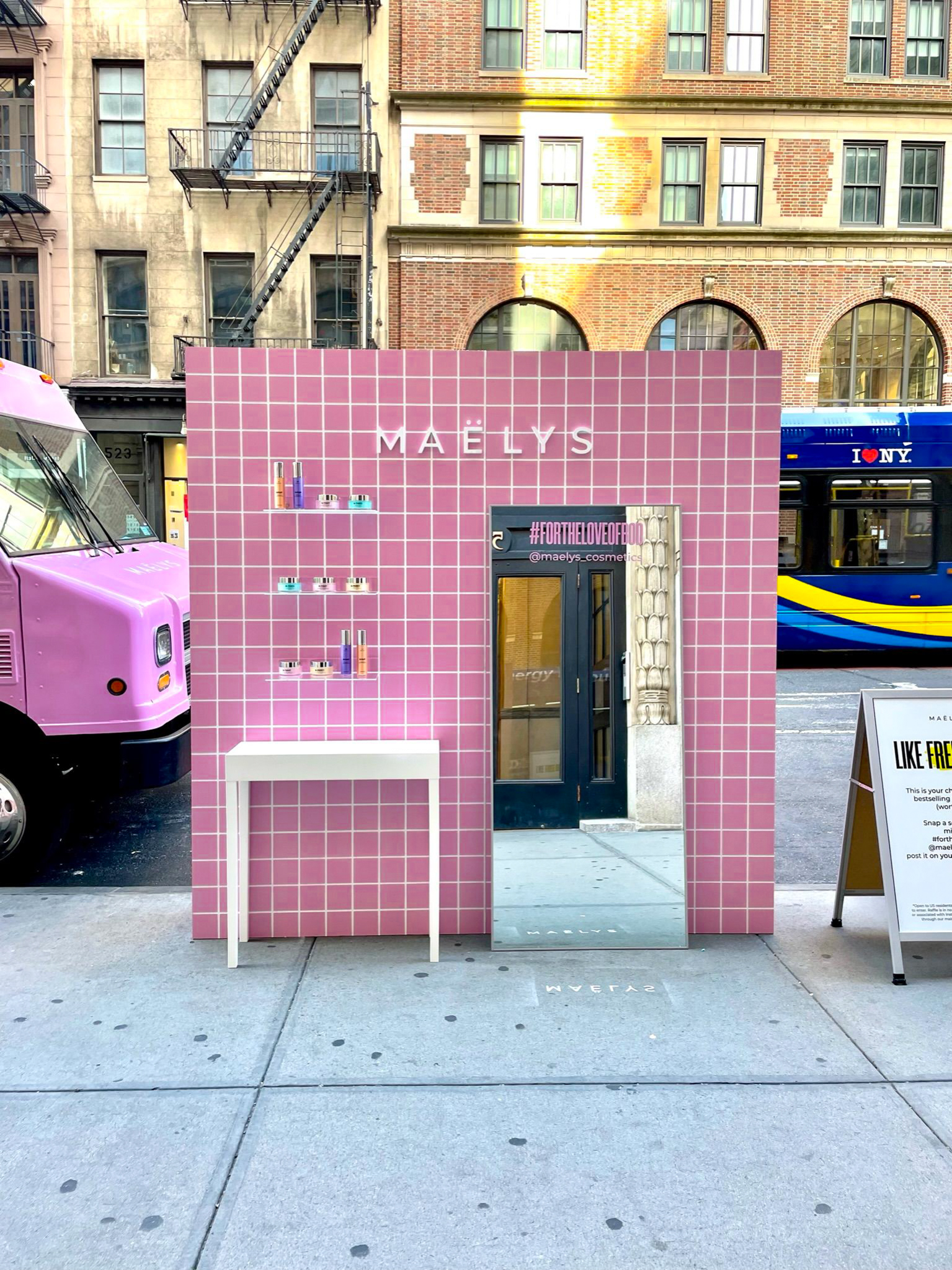 Pink House of Bod pop-up with MAËLYS sign and table on sidewalk; food truck, bus, buildings behind.