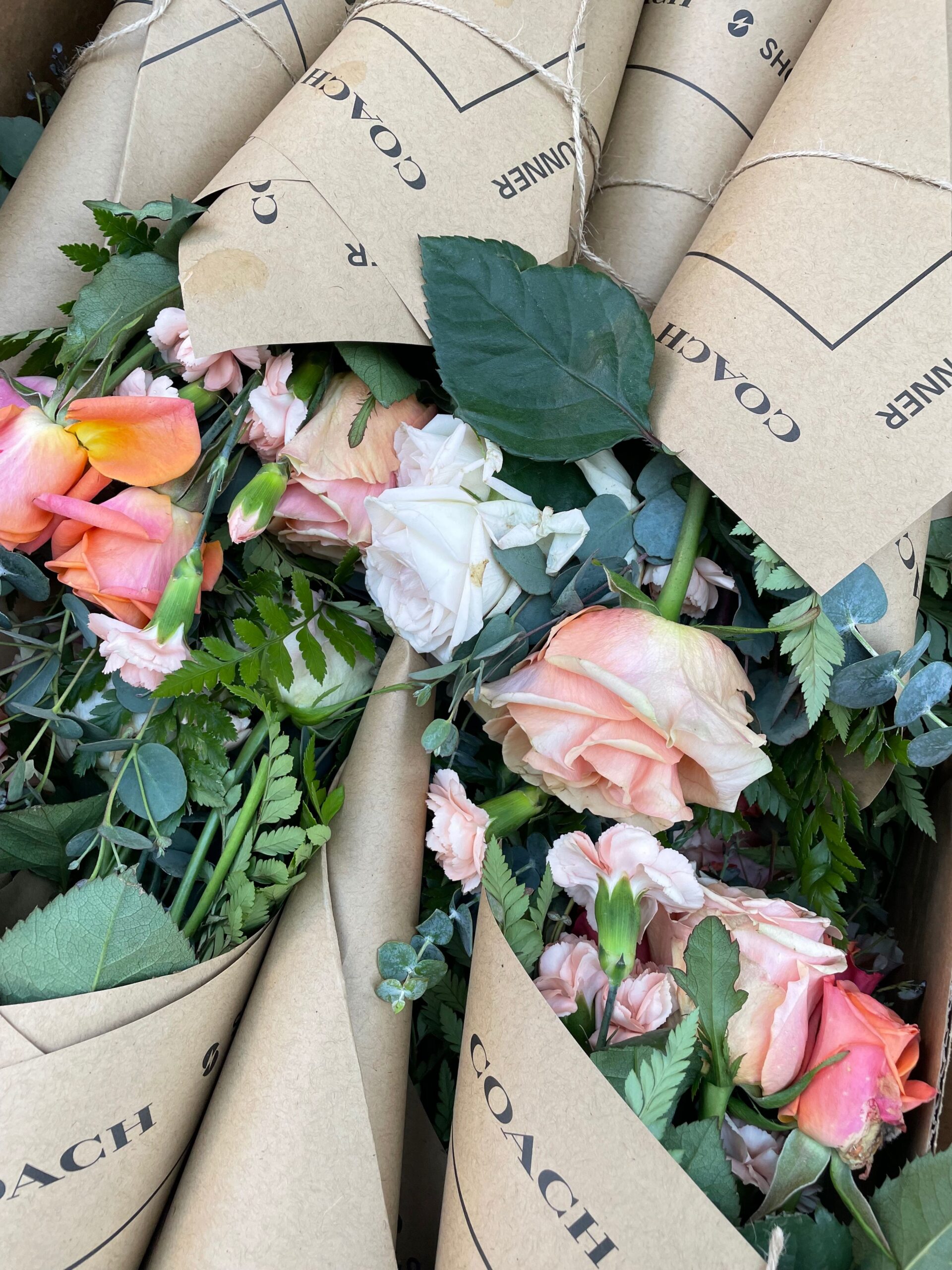 Pink, peach, and white rose bouquets with greenery wrapped in brown COACH paper, packed closely.