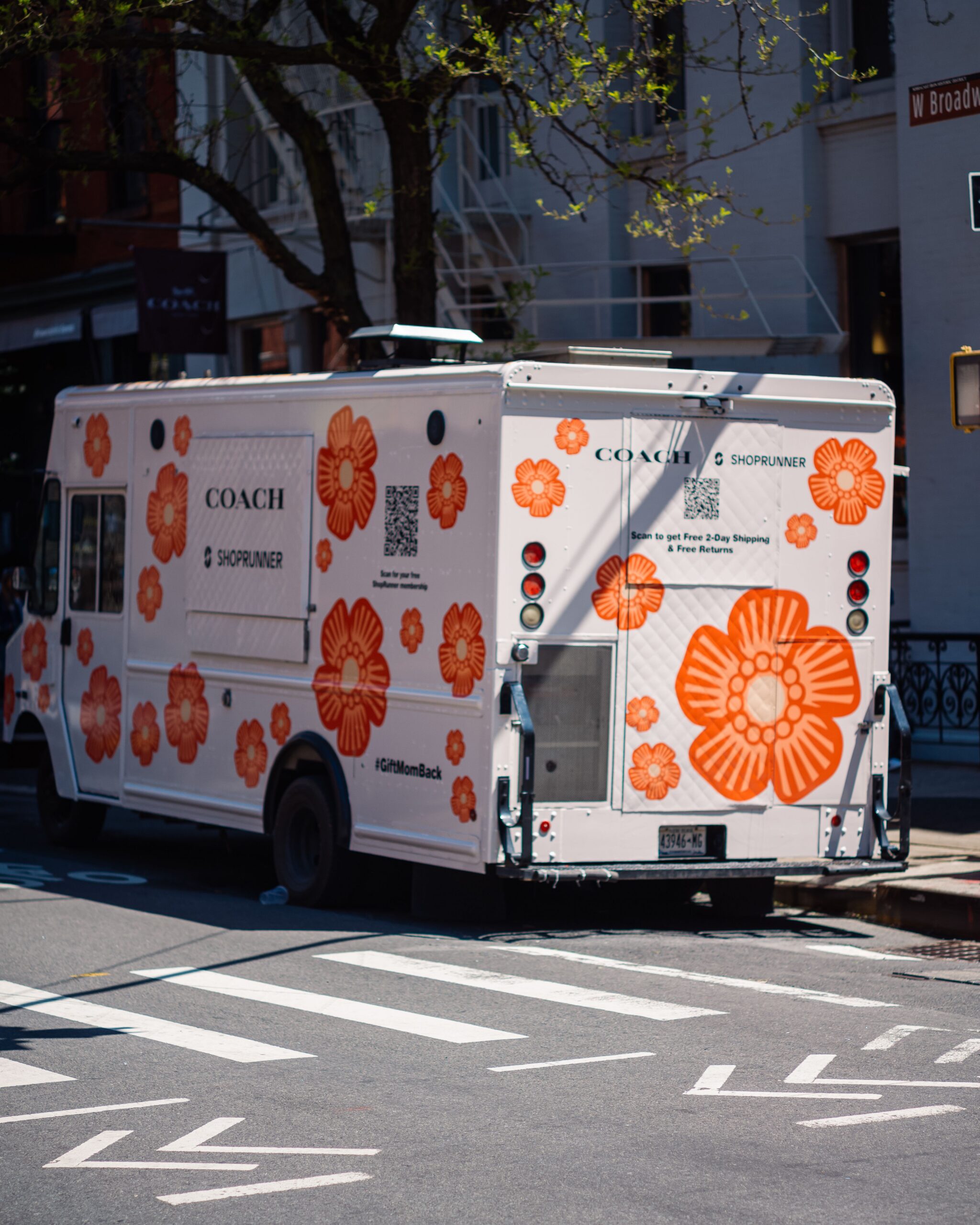 White delivery truck with orange floral designs and Coach branding parked by a city crosswalk.