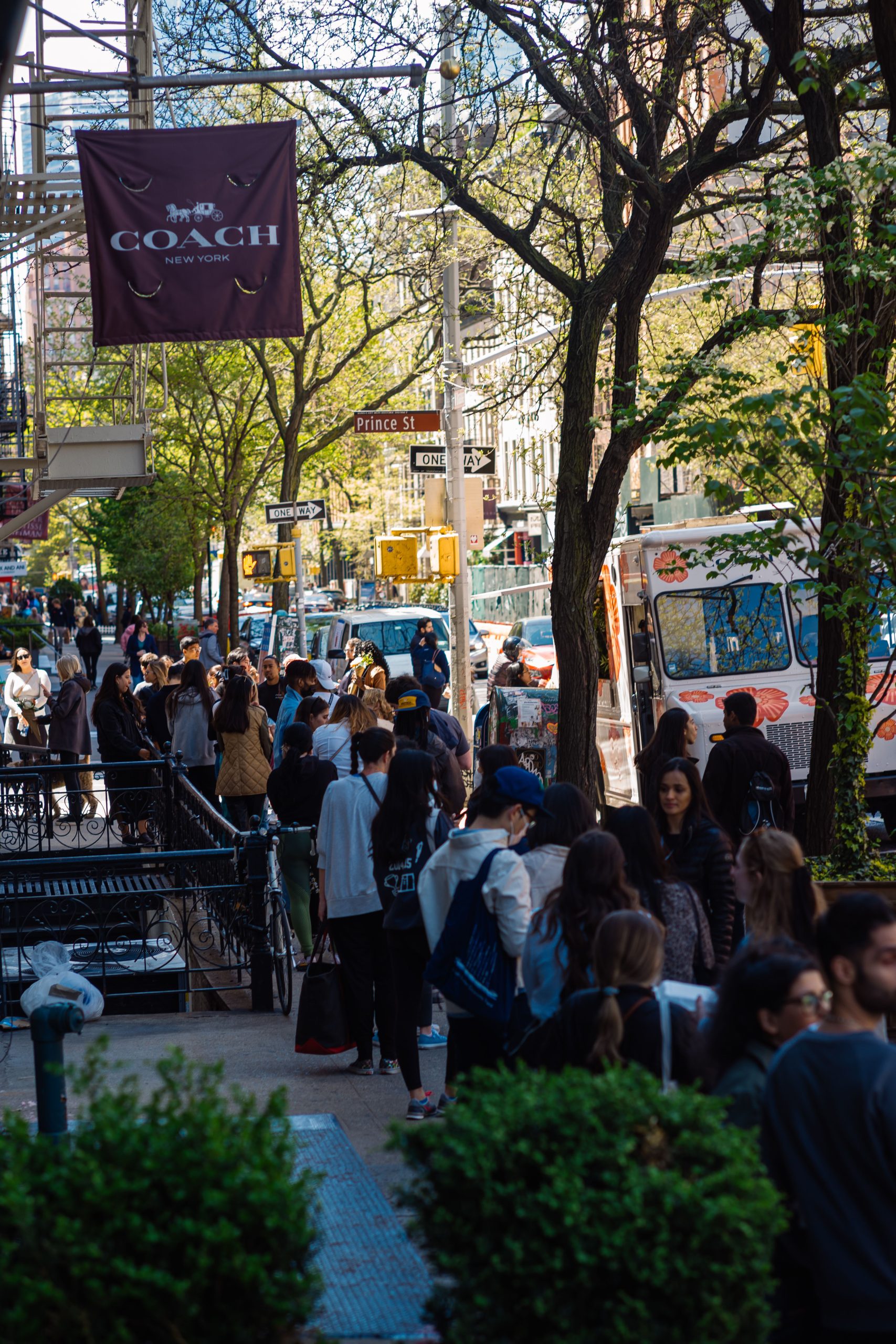 People line up by food trucks on a city sidewalk near a Coach Mother's Day pop-up banner.