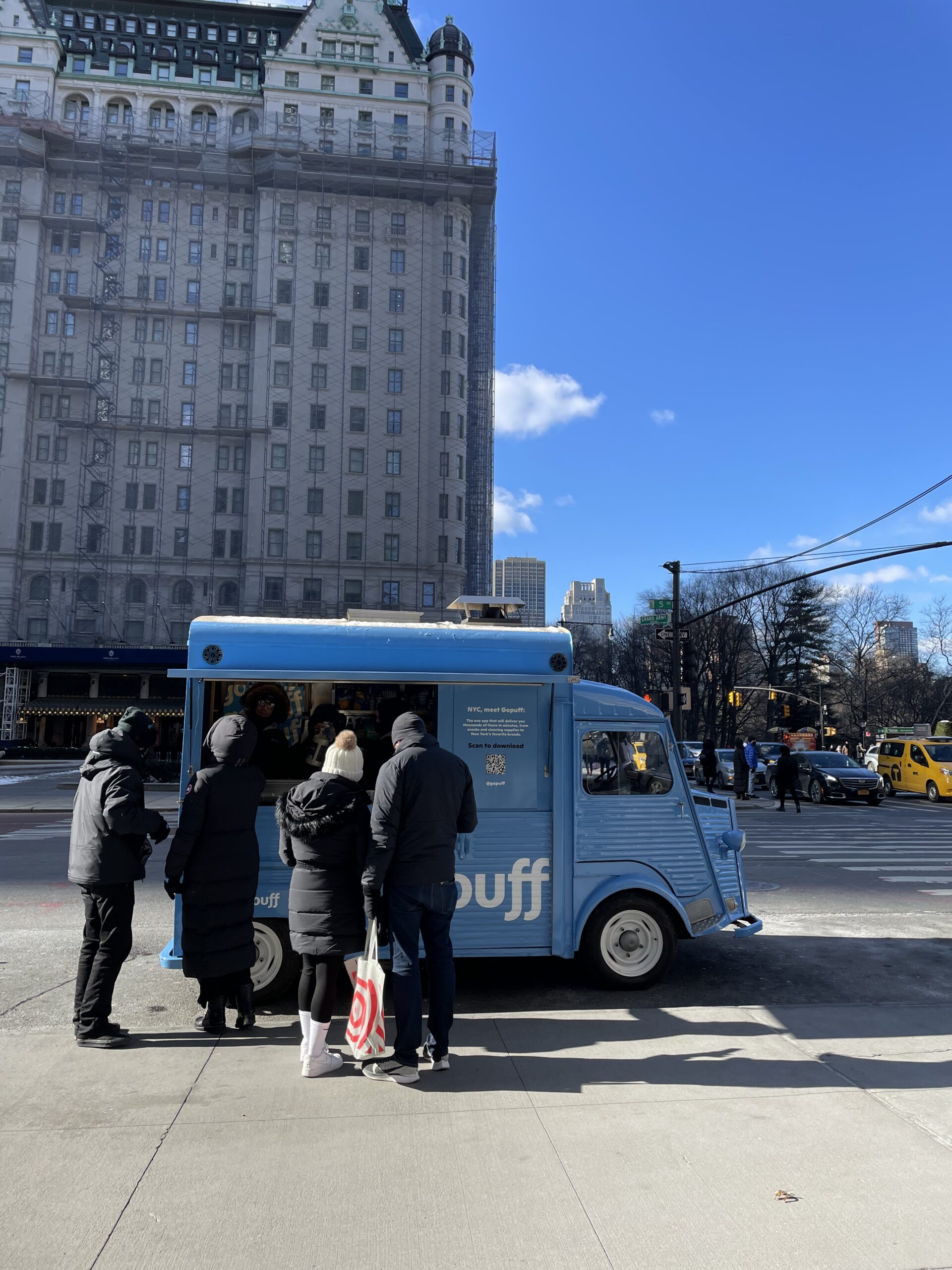 Blue food truck serves customers in winter coats; tall building and yellow taxis behind.