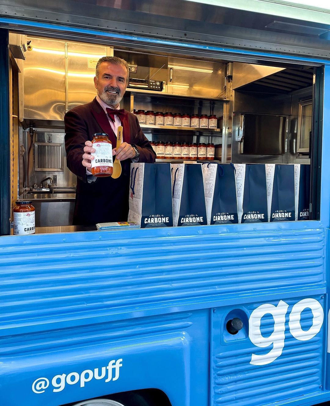 Man in suit holds Carbone pasta sauce jar inside blue food truck with "@gopuff" on the side.