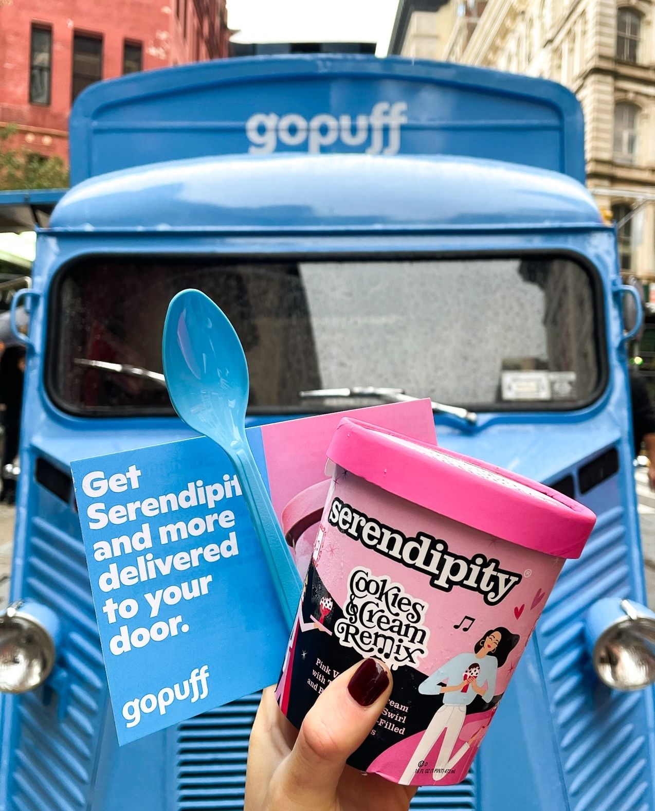 Hand holding Serendipity ice cream, blue spoon, and Gopuff flyer by a blue Gopuff truck on street.