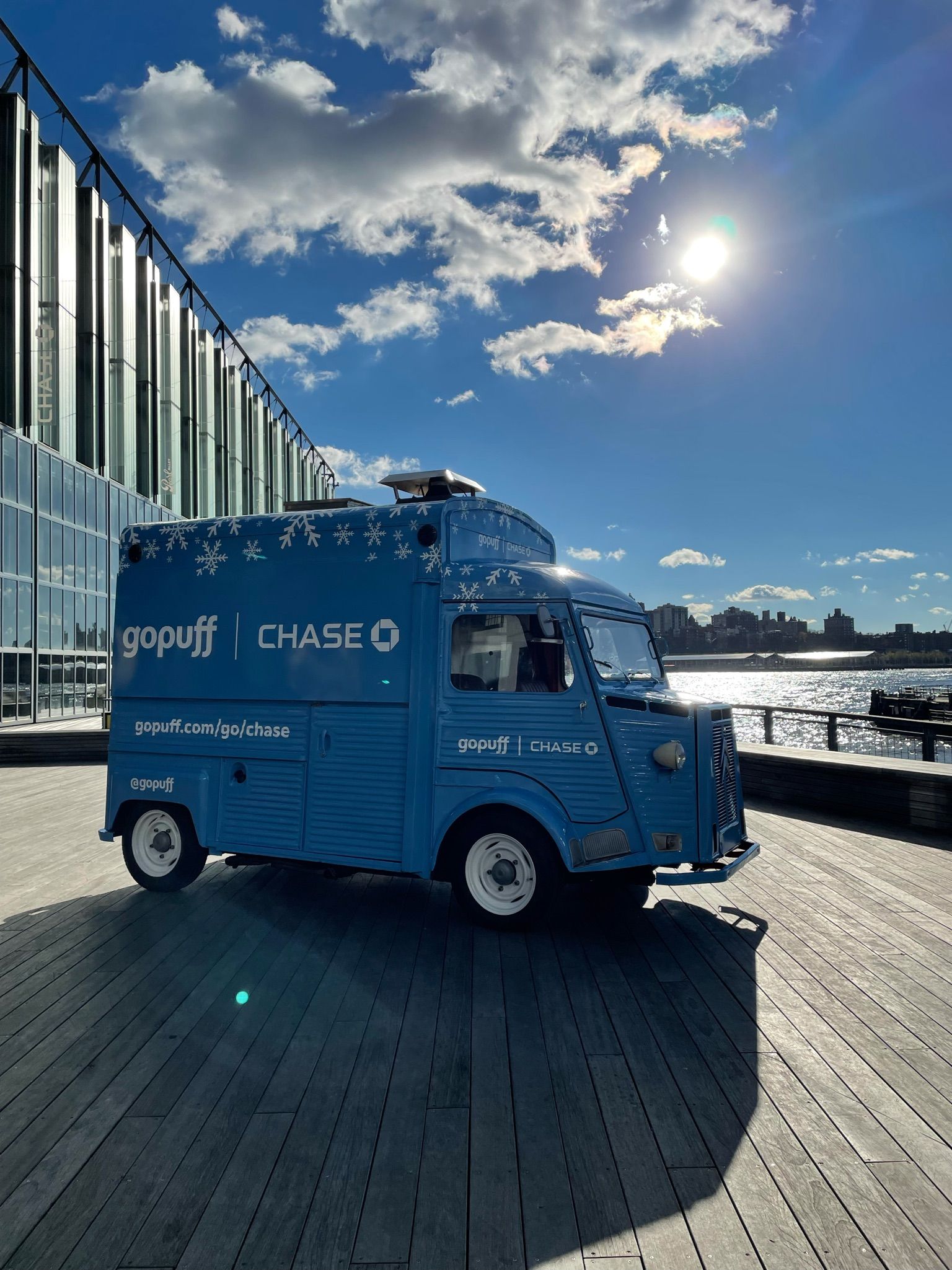 Blue Gopuff and Chase delivery van parked on a boardwalk by water with city skyline behind.
