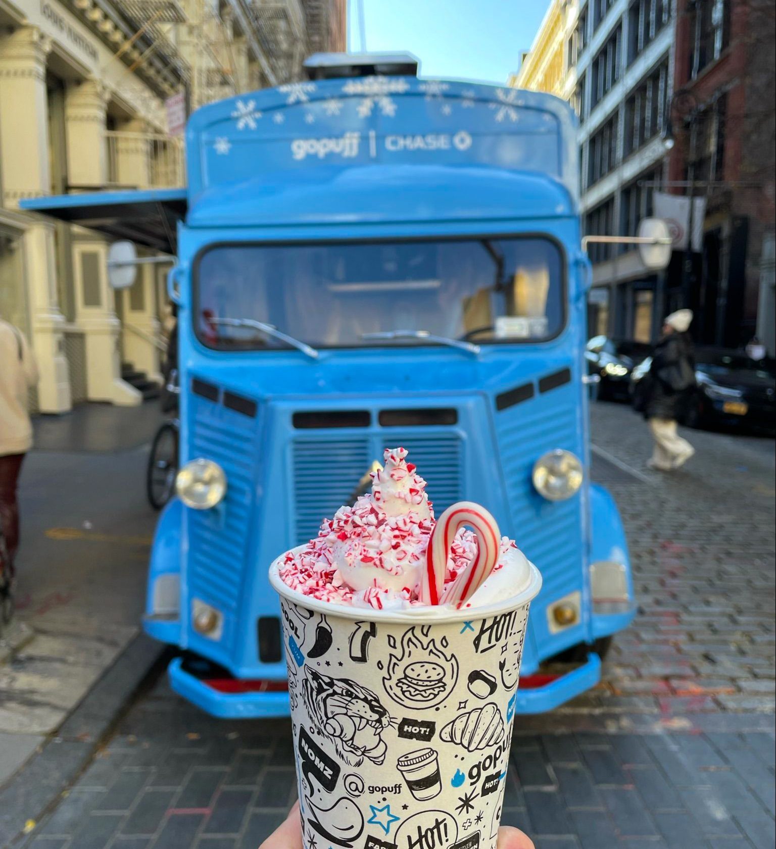 Hand holds hot drink with whipped cream and candy canes in front of a vintage blue food truck.