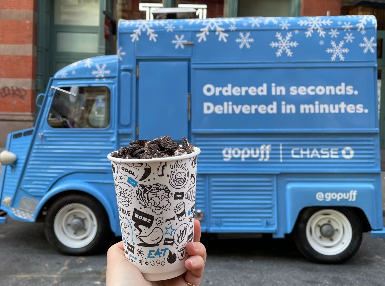 Hand holds cookie-topped cup in front of blue Gopuff truck with snowflake decorations and slogan.