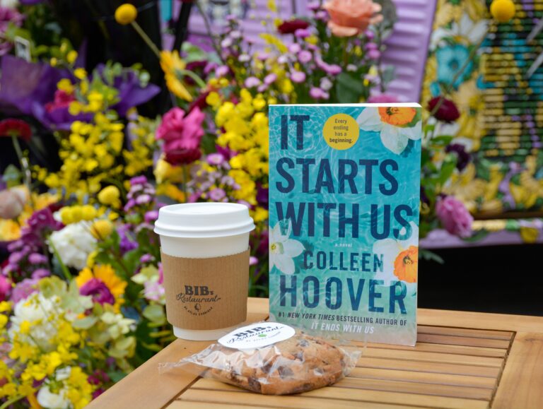 It Starts With Us" book next to a coffee cup and wrapped cookie, flowers behind them.