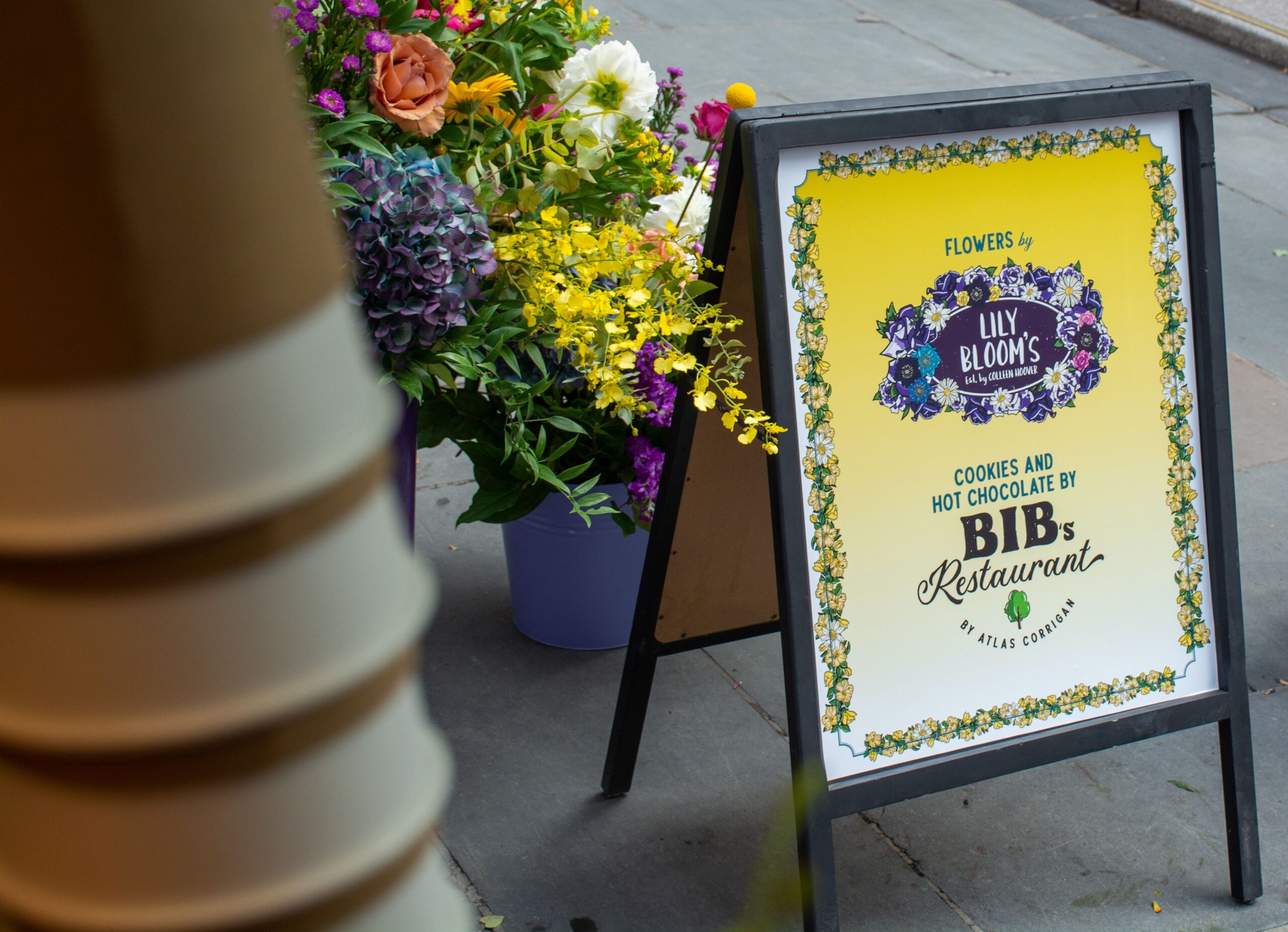Sidewalk sign for Lily Blooms flowers and Bib’s cookies with flower displays beside it.