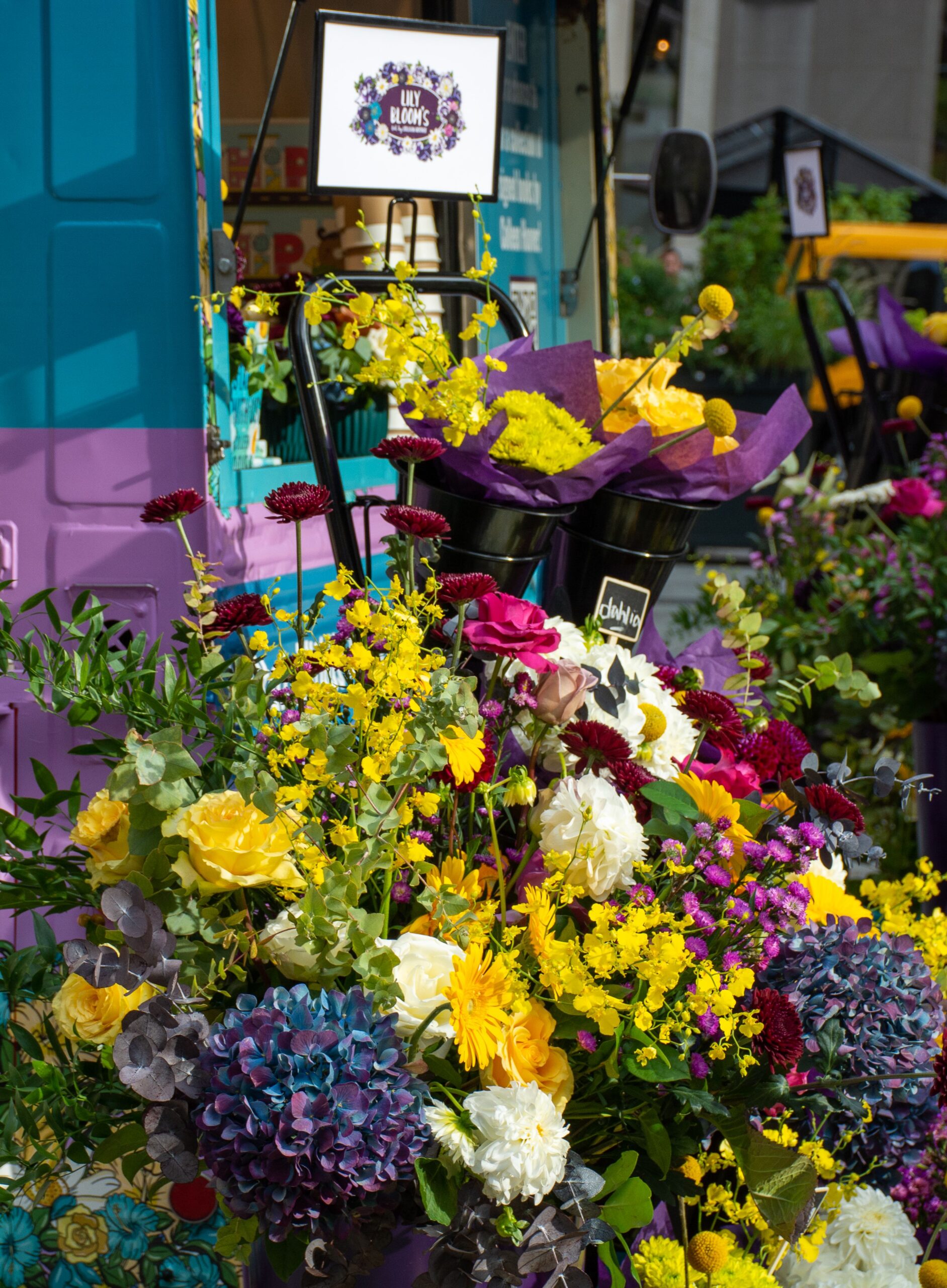Yellow roses, purple hydrangeas, and mixed flowers in front of a teal and purple Lily Blooms cart.
