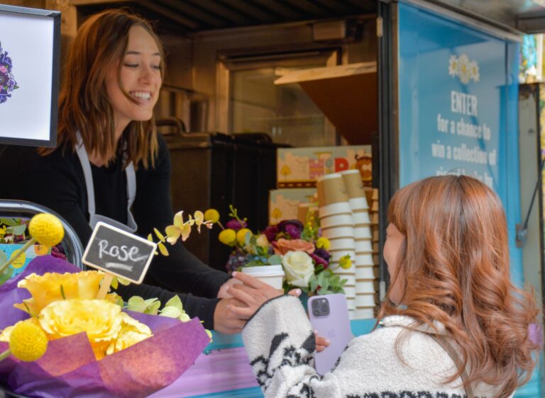 Woman in apron gives coffee to customer with phone at a flower-decorated food truck counter.