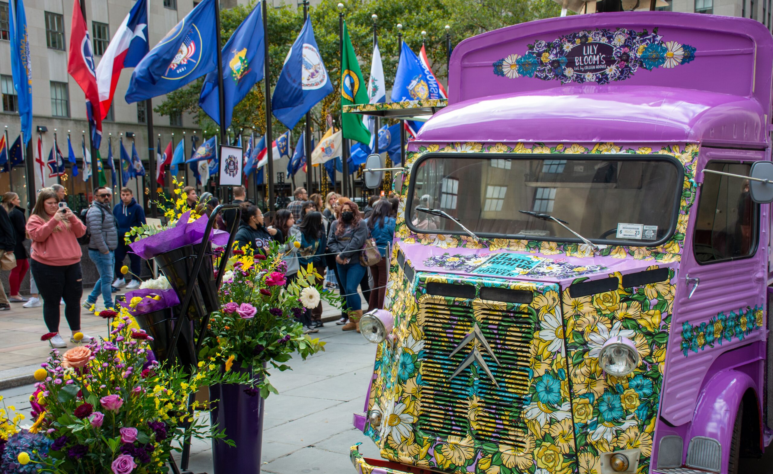Purple van with floral designs by flower displays as people take photos and wave flags in a plaza.