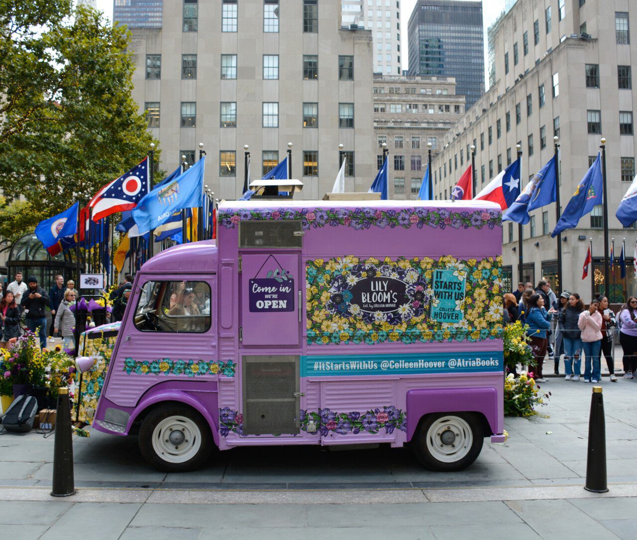 Purple Lily Blooms food truck with motto, parked by Rockefeller Center among people and flags.