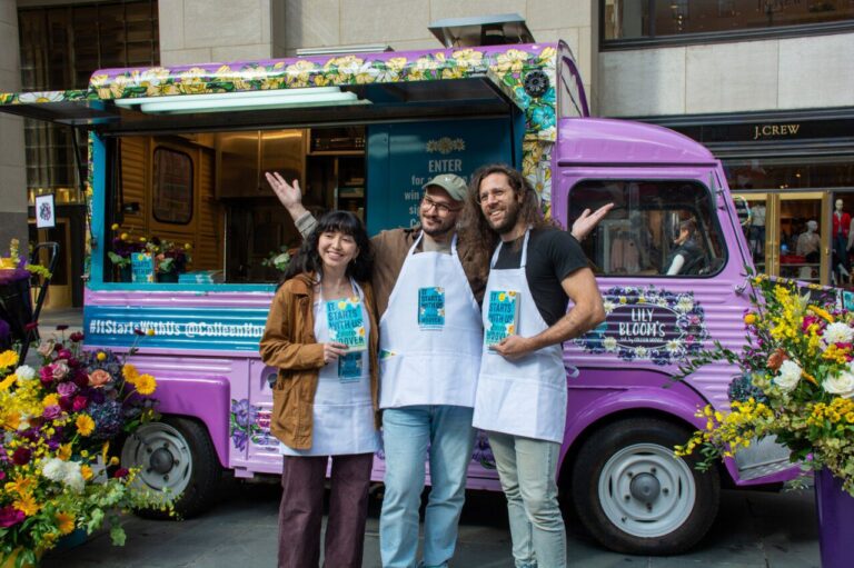 Three people in white aprons stand smiling by a purple flower-covered food truck, holding brochures.
