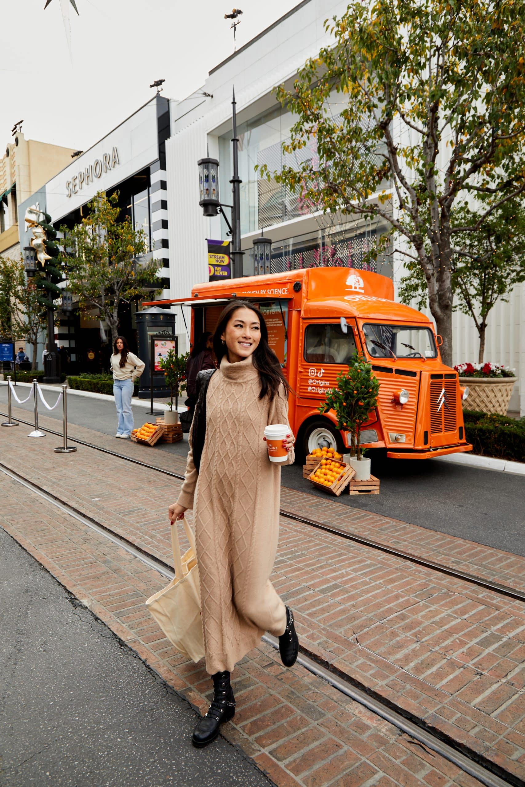 Woman in tan sweater dress and black boots holds coffee and tote, passing food truck near Sephora.