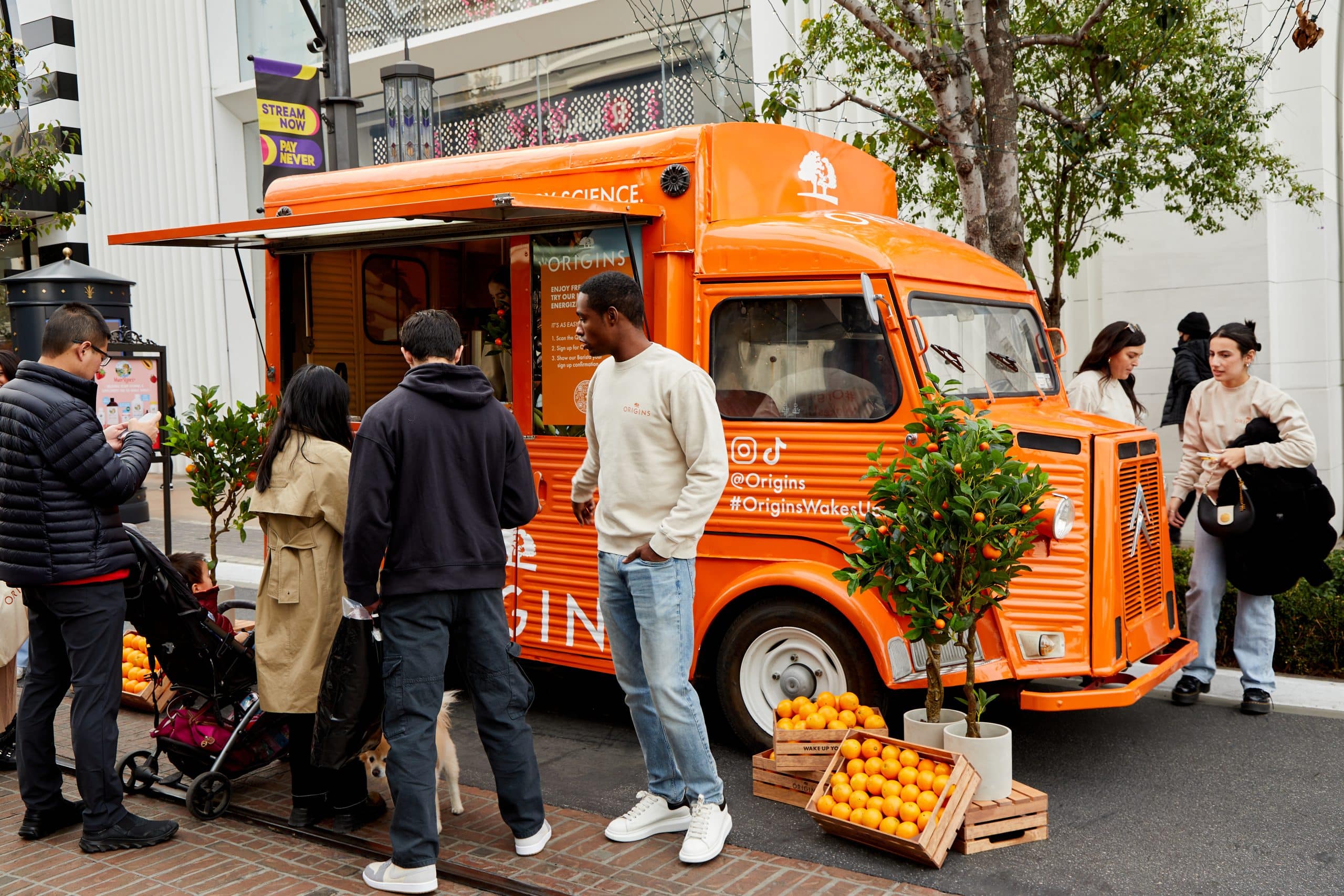 People gather at an orange Origins food truck with crates of oranges and small orange trees nearby.