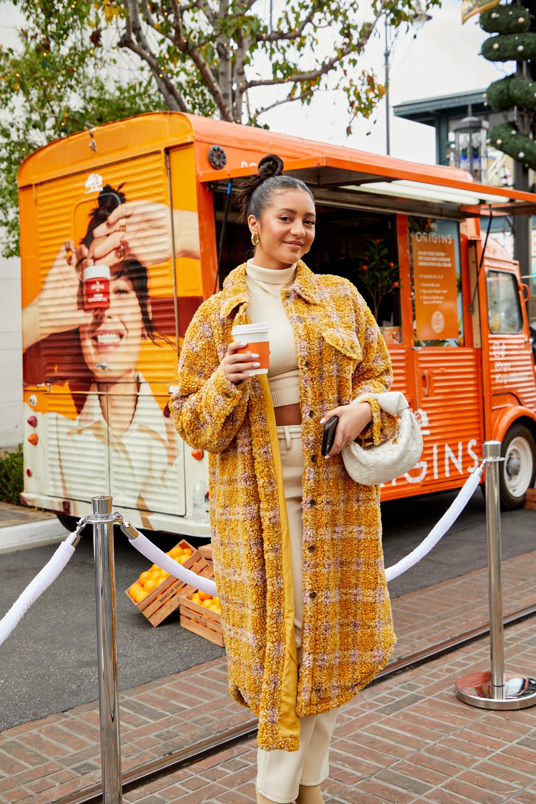 Woman in yellow plaid coat holds coffee and purse in front of orange food truck with oranges.