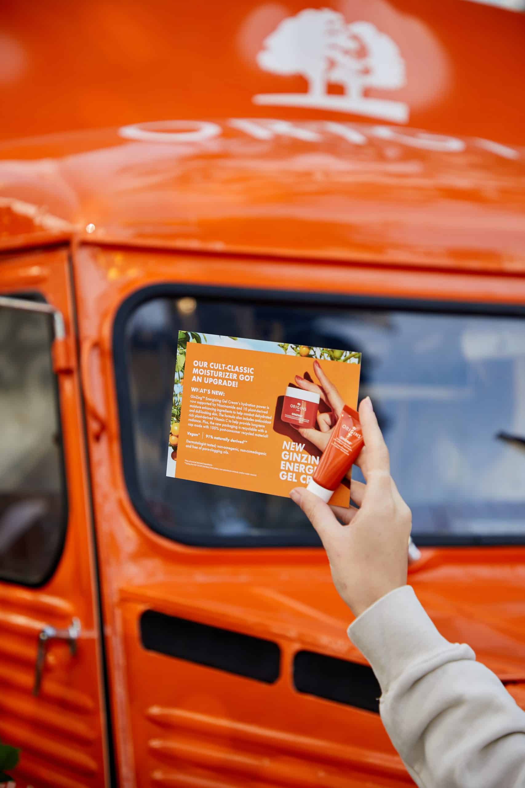 Hand holds a card and orange tube before an Origins truck; skincare info and logo visible.