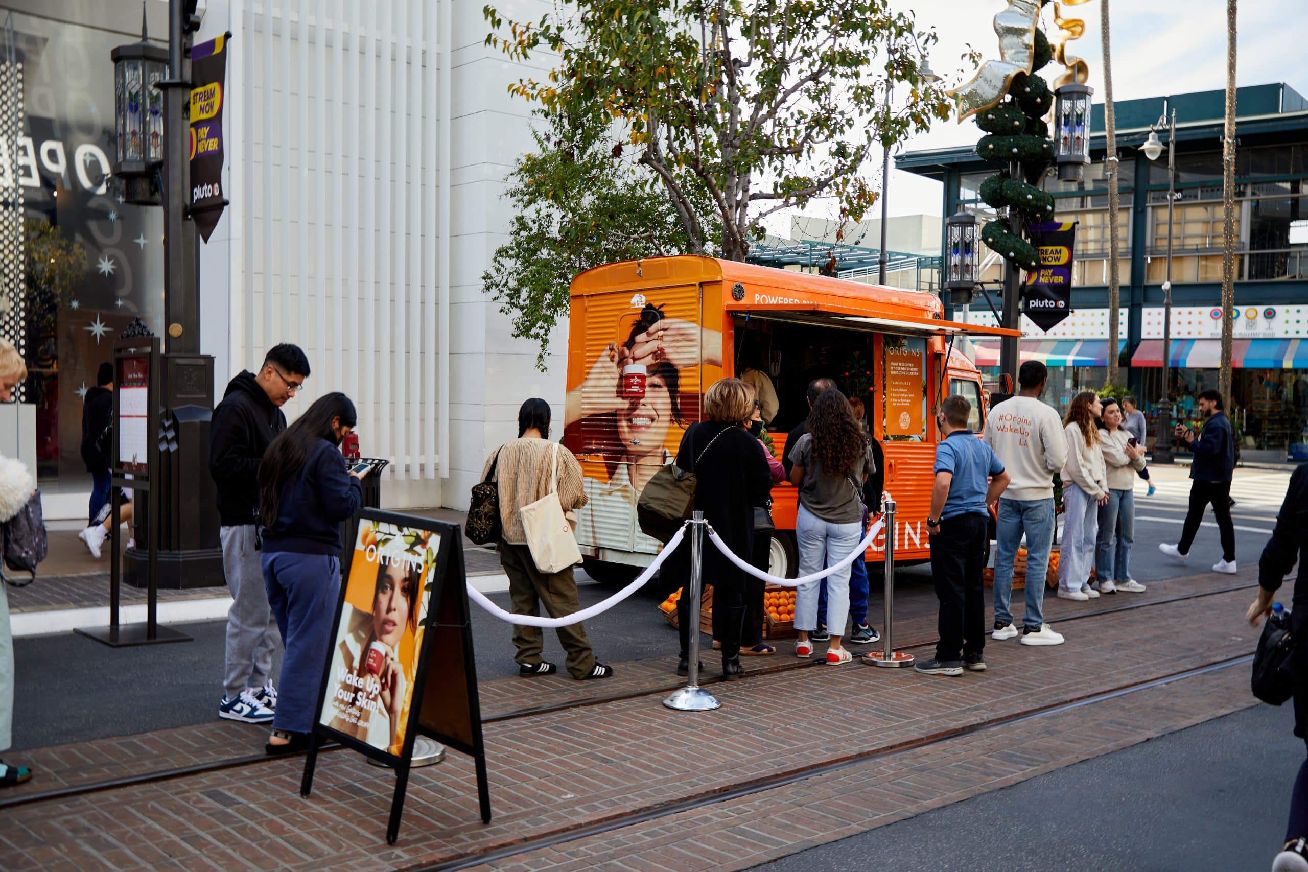 People line up at an orange pop-up food truck with a menu sign nearby on a city street.