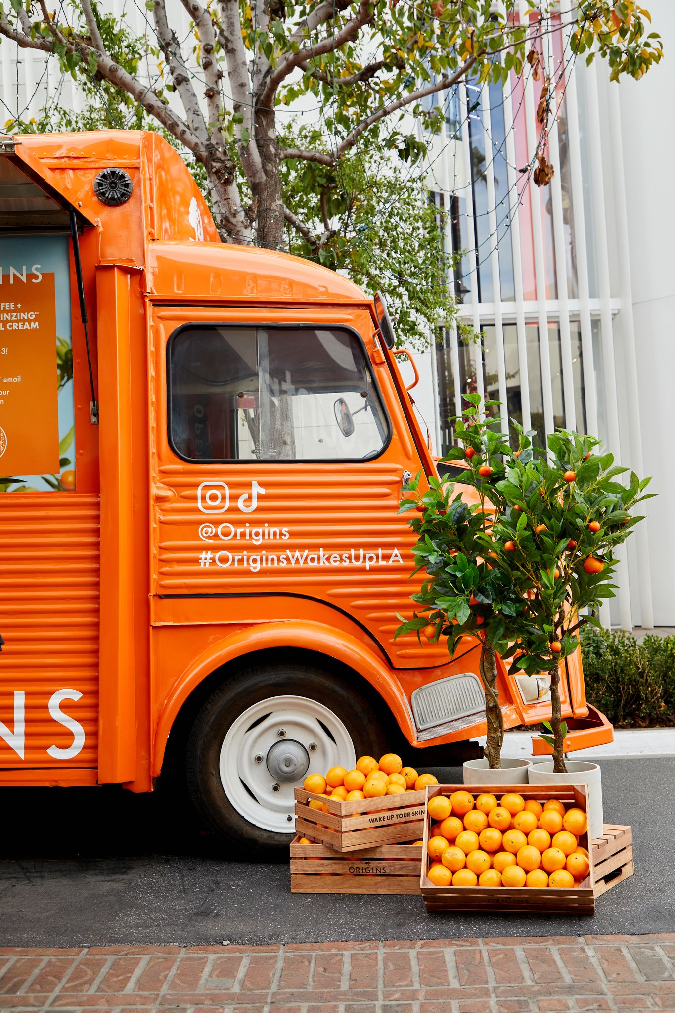 Orange food truck parked by crates of oranges and a tree; social media handles on the side.