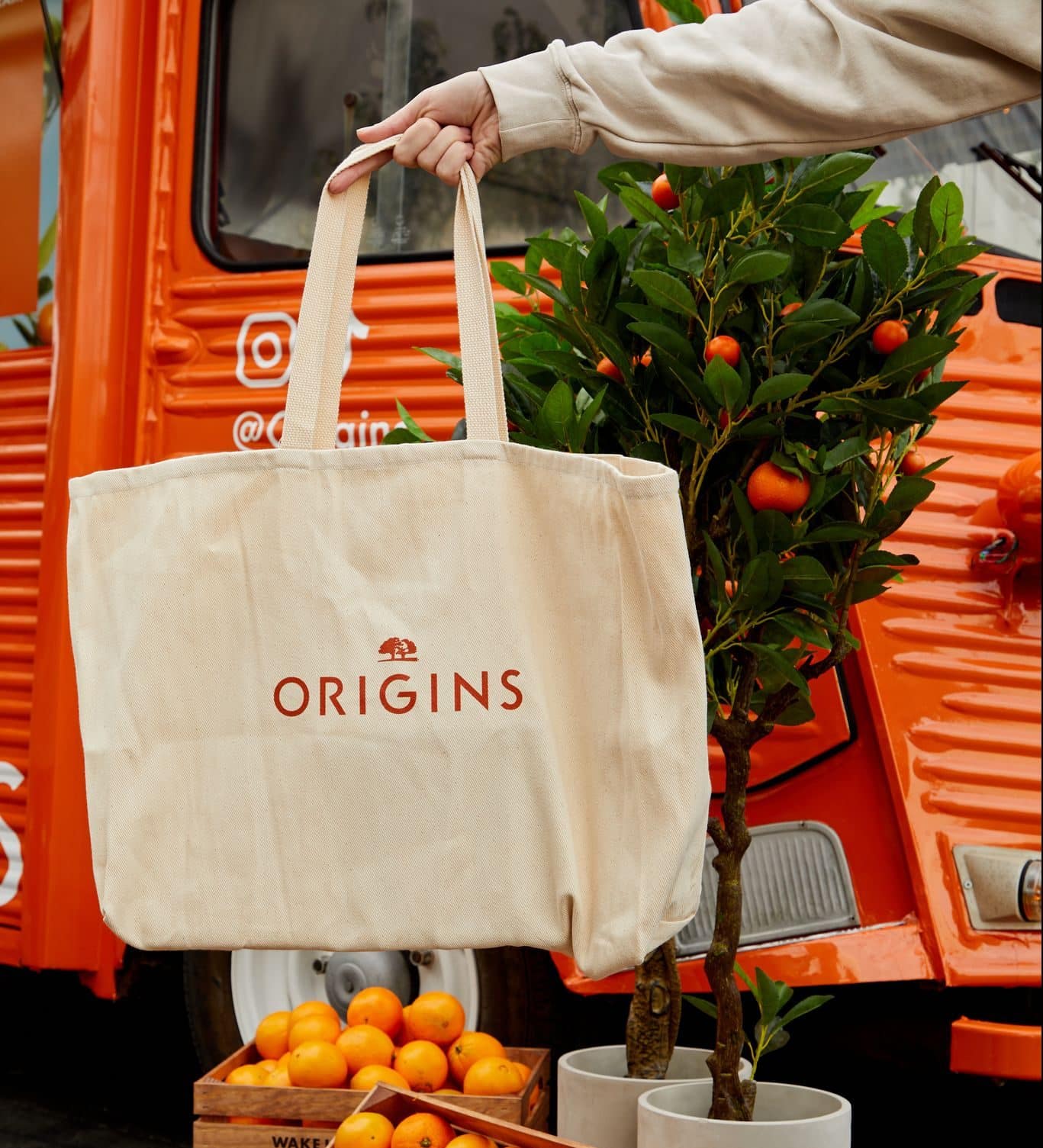 Person holding beige tote bag with ORIGINS text by orange food truck, oranges and trees nearby.