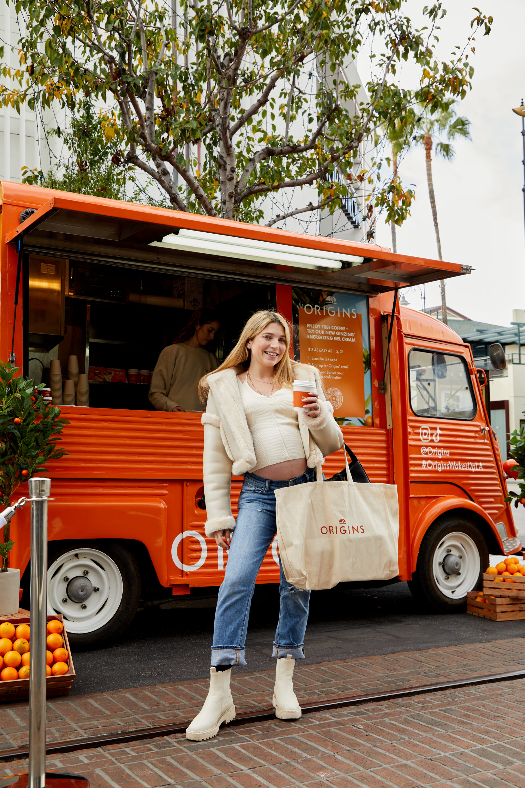 Woman with drink and Origins tote stands by orange food truck, oranges and tree nearby.