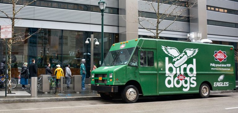 Green bird dog truck with wing graphics parked on city street as people with umbrellas gather nearby.