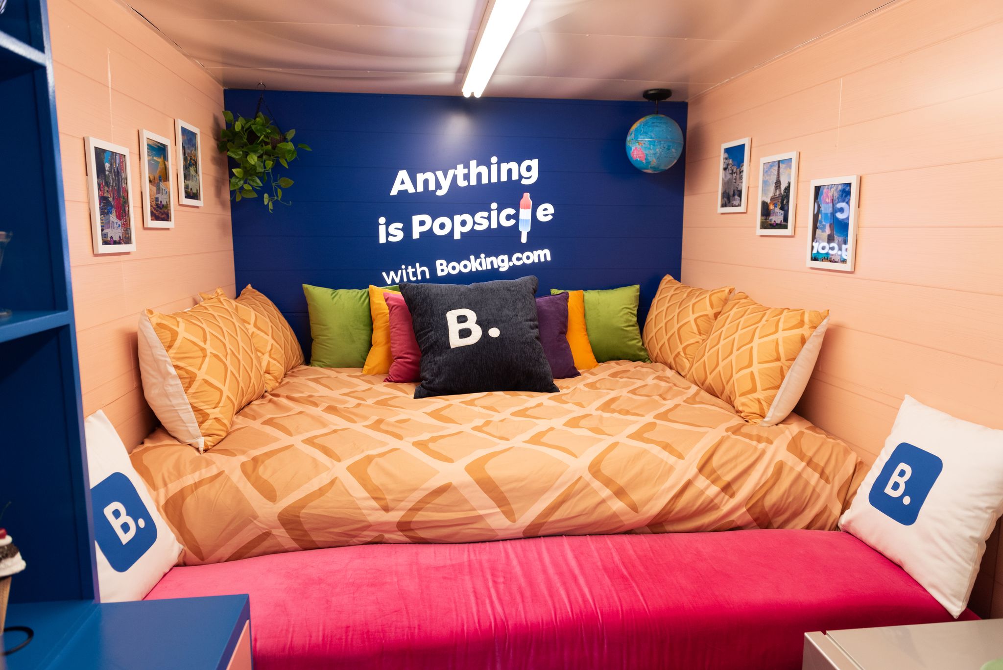 Room with a large bed, popsicle-patterned comforter, pillows, wall photos, plant, globe, and slogan.