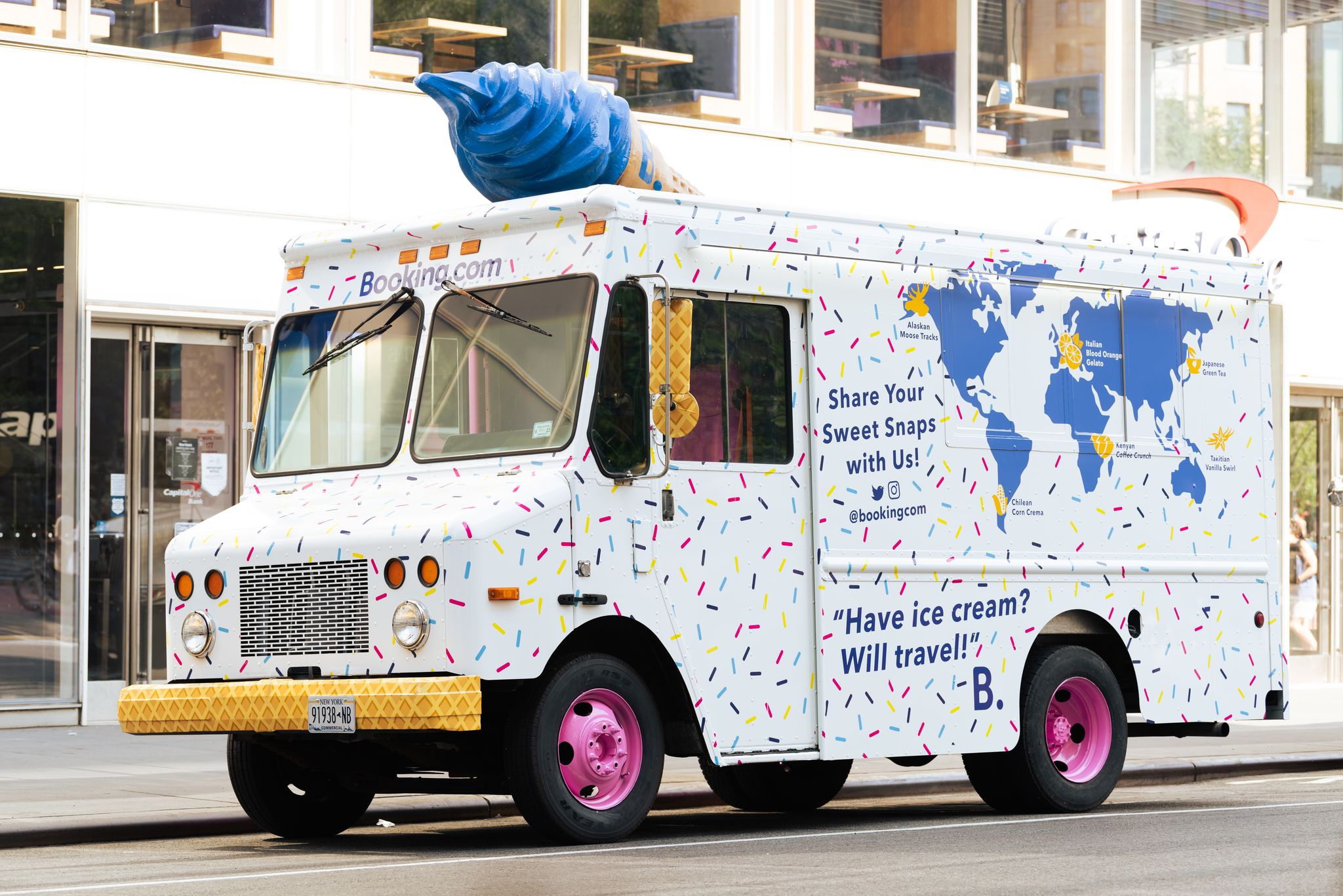 Booking.com ice cream truck with sprinkles, pink wheels, and big blue cone parked on city street.