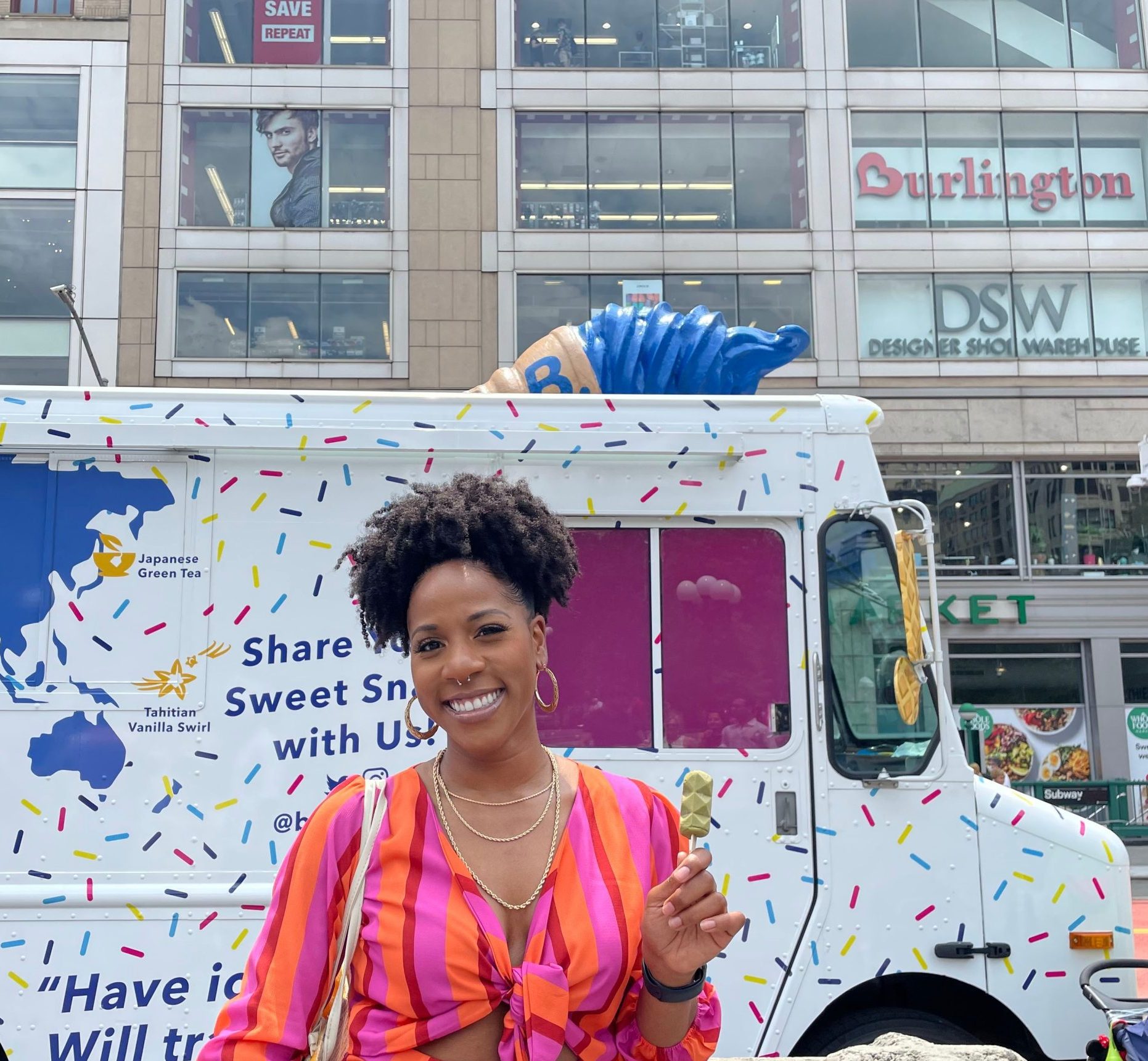 Woman holding ice cream cone by a sprinkle-decorated booking.com truck, city stores behind.